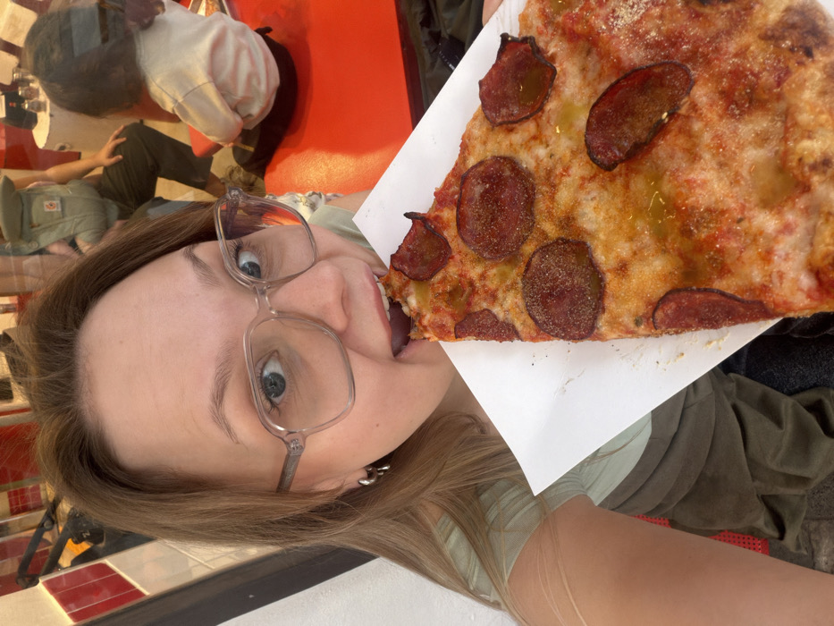 A candid, eye-level indoor photograph depicts a female subject actively consuming a slice of pizza within an eatery in Tel-Aviv, Israel. The primary figure, a light-skinned female in her 20s or 30s, possesses light brown hair and wears clear-framed eyeglasses and small silver hoop earrings. Her mouth is open, biting into a large, triangular slice of pepperoni pizza, which she holds with her left hand on a white paper surface. The pizza displays a cheese base, tomato sauce, numerous pepperoni slices, a visible oil drizzle, and dark seasoning. She wears a light green top and directly faces the camera with wide eyes.
The background reveals an interior space, likely a reflection, featuring a prominent bright red table or counter. A small child with dark hair, dressed in a light-colored long-sleeved top and dark pants, sits at this red surface, facing away from the camera. A partially visible adult figure, also oriented away or to the side, is present behind the child, wearing dark pants and a beige baby carrier. Architectural elements featuring red and white horizontal stripes are visible, consistent with commercial decor, and small condiment shakers are positioned on the red surface.