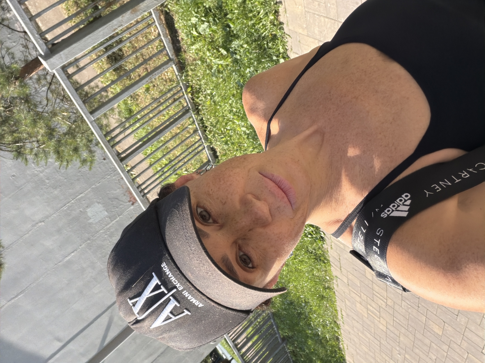 A woman wearing a black Armani Exchange baseball cap and a black tank top takes a selfie outdoors in an urban environment. She has brown eyes and freckles on her face and chest. A black strap with the Adidas logo and the word "STELLA" is visible across her shoulder. She is positioned in front of a grey wall and a metal fence, with green grass and a paved walkway in the background. The lighting suggests it is daytime.
