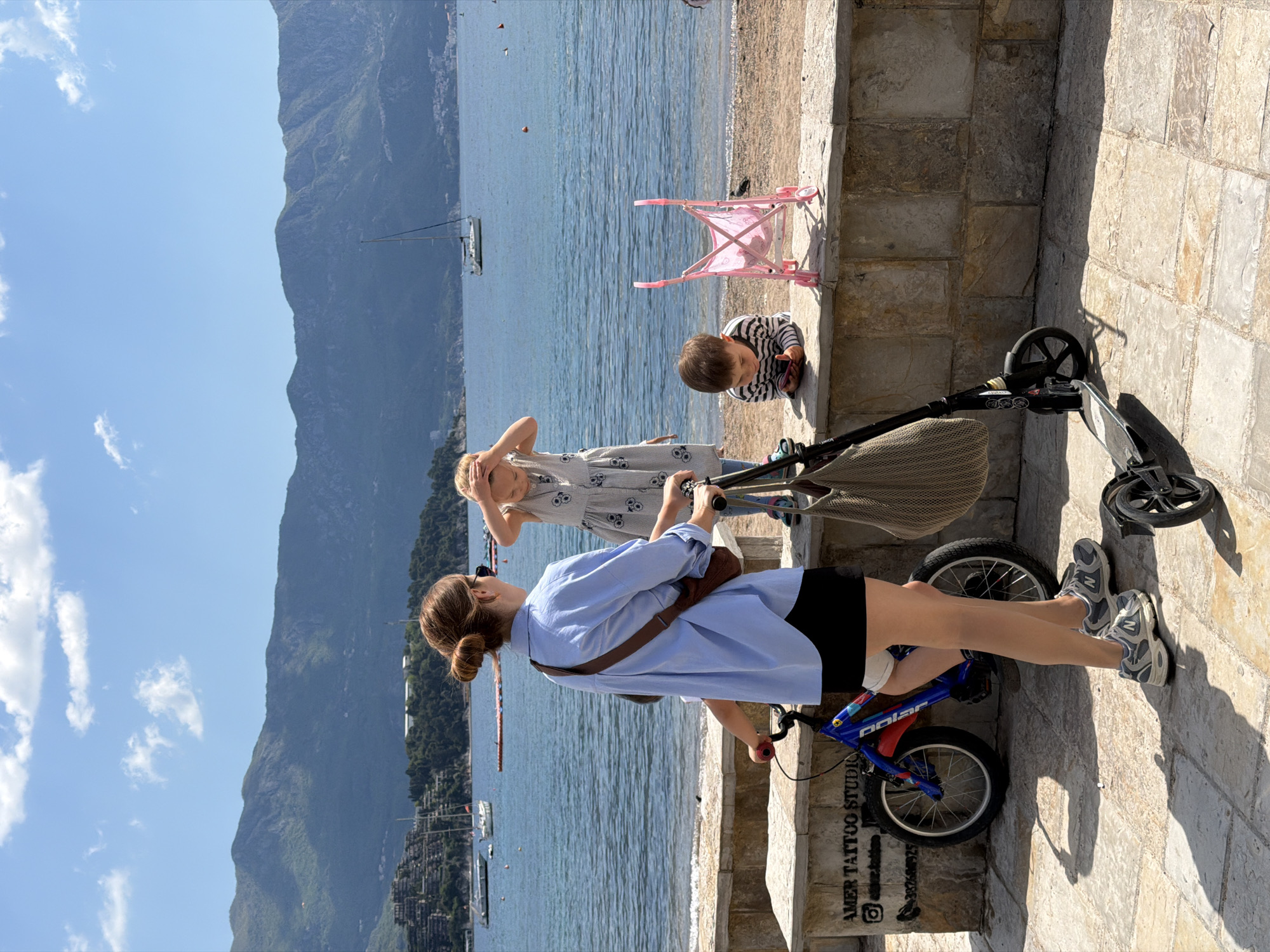 A medium outdoor shot depicts a woman on a child's bicycle on a stone staircase in Budva, Montenegro. She is turned away from the camera, wearing a light blue long-sleeved shirt, black shorts, and gray sneakers, with her brown hair in a bun. A brown bag is slung across her shoulder. To her left are a young girl and boy, standing on the edge of a stone wall overlooking a sandy beach and the blue Adriatic Sea. The girl has blonde hair, a light-colored dress with a skull pattern, and her hands near her head. The boy is wearing a striped shirt and is seated near a pink toy stroller. Further to the right is a stroller and scooter. In the background are mountains, the sky, and a couple of sailboats. The environment is sunny and bright. A tattoo studio advertisement is visible on the stone at the bottom right.