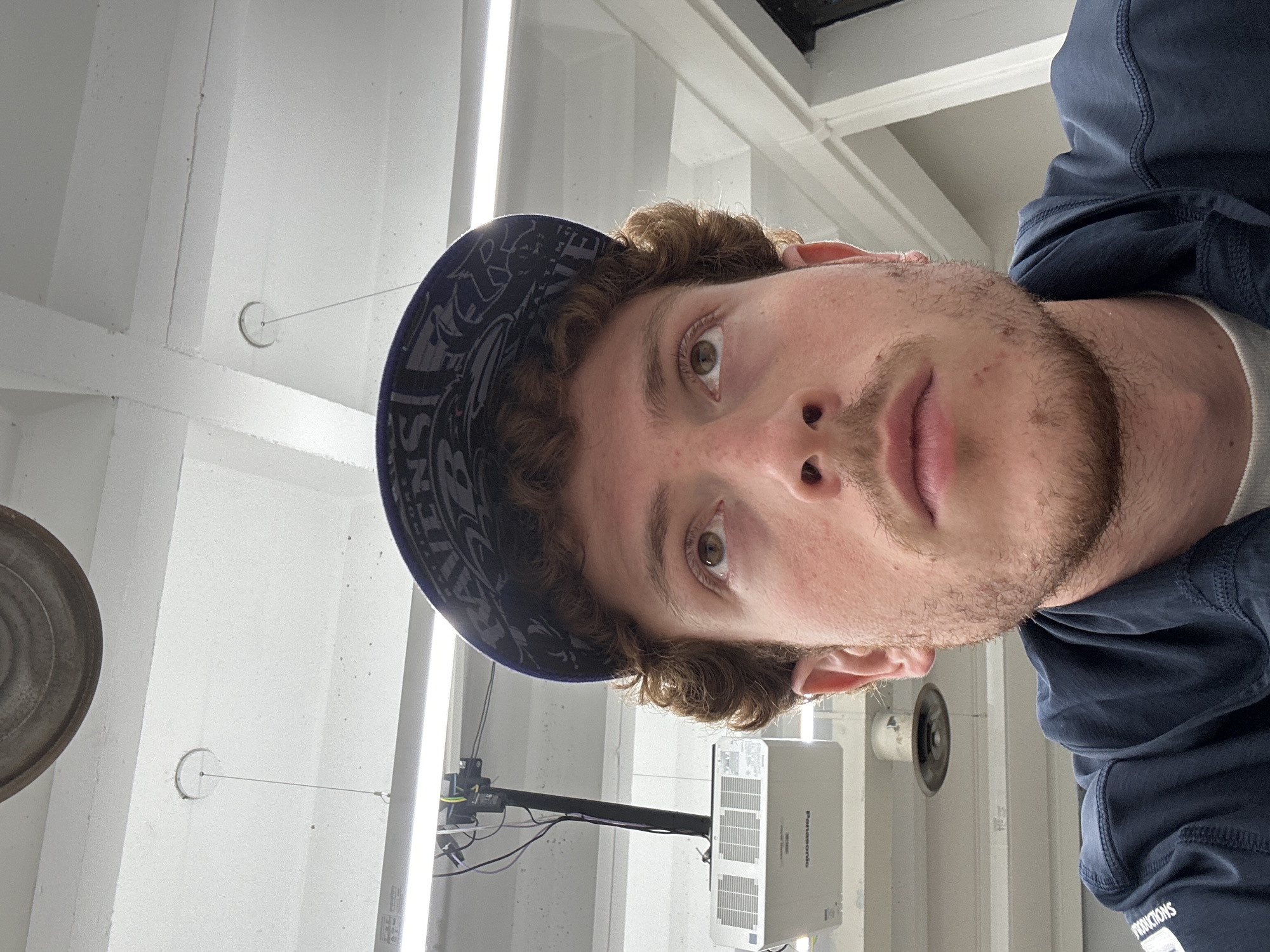 This close-up image, originally rotated on its side, captures a young man looking directly at the camera with a neutral expression. He has light brown, curly hair, stubble or a short beard, and light brown eyes. He is wearing a dark blue baseball cap featuring a "RAVENS" logo, indicative of the Baltimore Ravens sports team. Over a light-colored undershirt, he wears a dark blue hooded top, on which the word "PRODUCTIONS" is visible near his right shoulder.
The background depicts an indoor environment with a white or light grey ceiling and walls, suggesting a concrete structure. Bright, linear artificial lights illuminate the space. A white "Panasonic" brand projector is mounted from the ceiling on a black arm, with several cables visible around its base. To the left, a portion of a metallic, circular object is visible, possibly a weight or part of a fixture. To the right, another circular object, potentially a vent or speaker, is seen. A thin wire or string also hangs from the ceiling, attached to a small circular fixture. The setting appears to be a functional, casual space such as a studio, workshop, or classroom, located in New York, United States of America. The time of day and weather are not discernible.