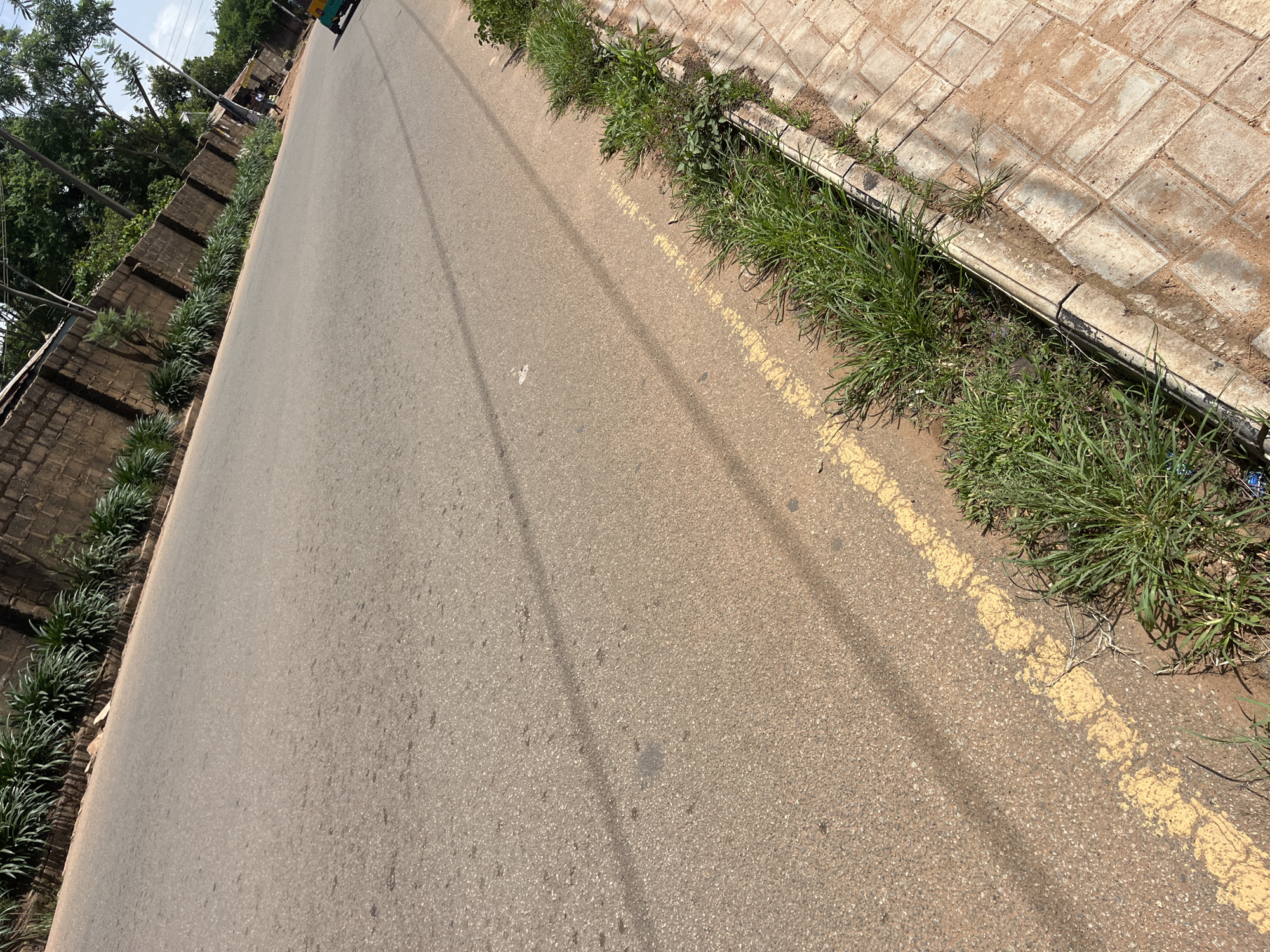The image shows a street scene in Gura Top, Nigeria, captured from a high angle, likely from a vehicle. The road, paved with asphalt, dominates the foreground, with a dashed yellow line marking the lane. To the left, a low brick wall runs alongside the road, topped with neatly planted, spiky green plants. Beyond the wall, lush green trees and foliage are visible, indicating a well-vegetated area. On the right side of the road, a paved sidewalk or walkway made of interlocking bricks is present, bordered by a concrete curb. Thick green grass grows along the edge of the road and sidewalk, some of which is encroaching onto the paved areas.
The scene appears to be set during the daytime, with clear shadows cast on the road, suggesting sunny weather. The overall impression is that of a well-maintained urban or suburban street in a tropical or subtropical climate. No people are clearly visible, but a hint of a vehicle, possibly a yellow and green truck or bus, is present in the upper right corner, suggesting traffic. The lighting and the presence of green vegetation suggest it might be morning or afternoon. No visible text is present in the image.