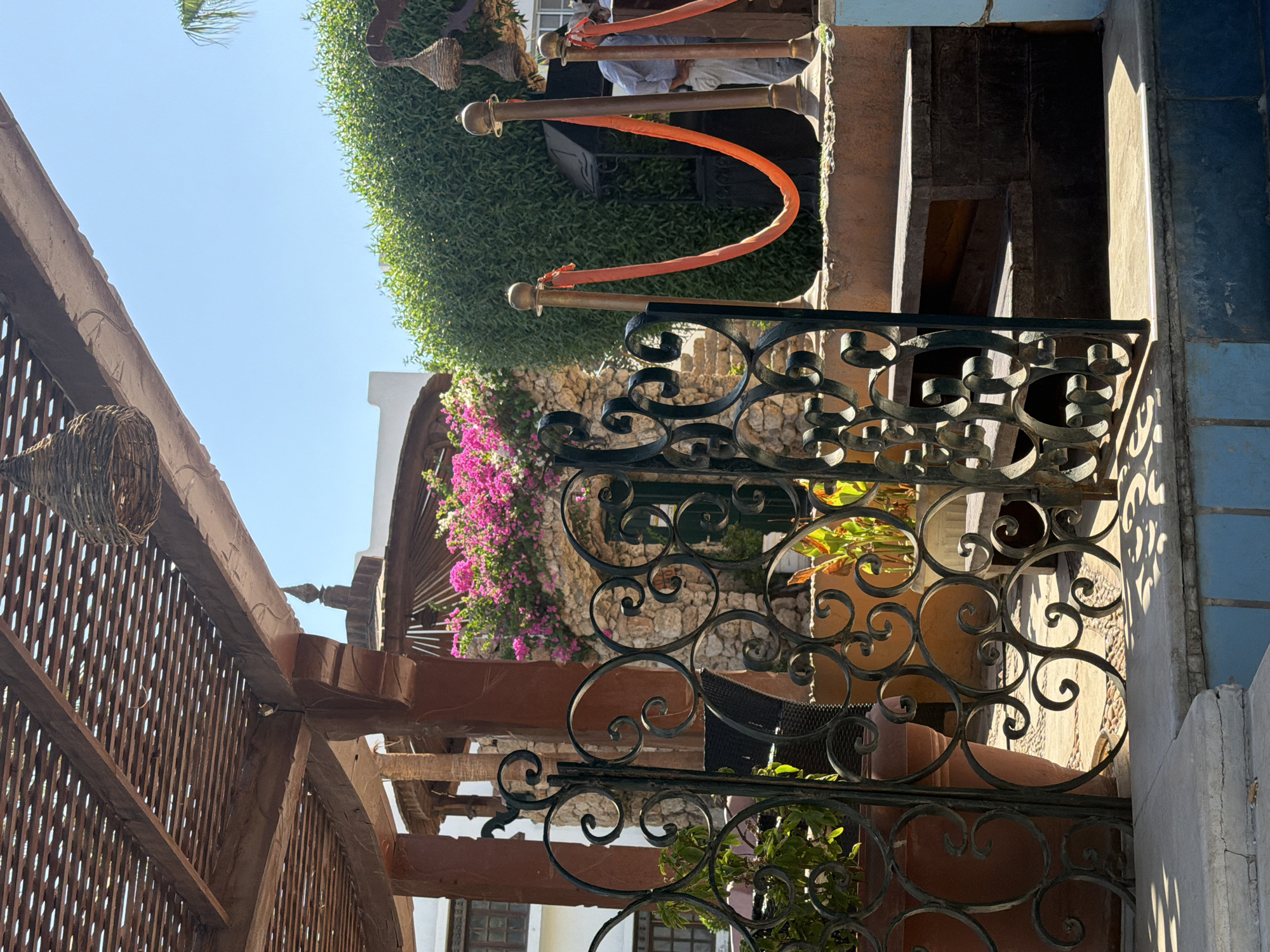 The image depicts an outdoor scene in Sharm El Sheikh, Egypt, likely a resort or restaurant area. A decorative wrought-iron gate with swirling patterns is in the foreground, partially open, revealing a glimpse of plants in large terracotta pots. Beyond the gate, a stone wall is adorned with vibrant pink bougainvillea flowers. To the right, a rope barrier with orange fabric separates an area, with a man in a light-colored shirt and trousers standing near a green hedge. Wooden steps lead up to a raised platform. The scene is bathed in bright sunlight, casting sharp shadows, indicating it is daytime. The overall impression is one of a lush, inviting outdoor space in a warm climate.