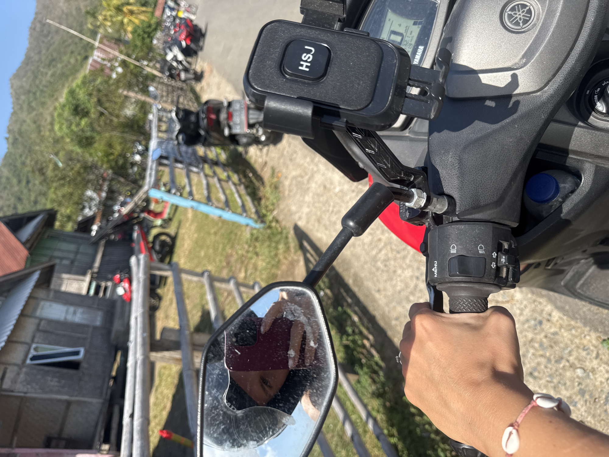 A first-person perspective image shows a hand on the handlebar of a red and black Yamaha scooter. The hand is wearing a bracelet made of pink string and white seashells. A phone mount with the letters "HSJ" is attached to the handlebar. In the reflection of the scooter's mirror, a person wearing a black helmet and a maroon shirt is visible, holding a red smartphone. The scooter is parked on a paved road in a village setting, with a wooden fence and simple houses in the background. The environment suggests a sunny day with a hilly, vegetated landscape in the distance. The specific location is the village of Kertasari, Indonesia.