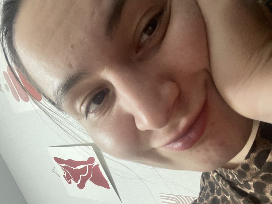 A selfie shows a fair-skinned woman with brown hair, eyes, and lips. She's tilted her head, resting a hand on her cheek. Her facial expression is warm and slightly smiling. The background includes a white wall adorned with abstract art, one piece in red. The woman is wearing a leopard-print top with a white edge visible. The camera angle gives a close-up view of her face.
