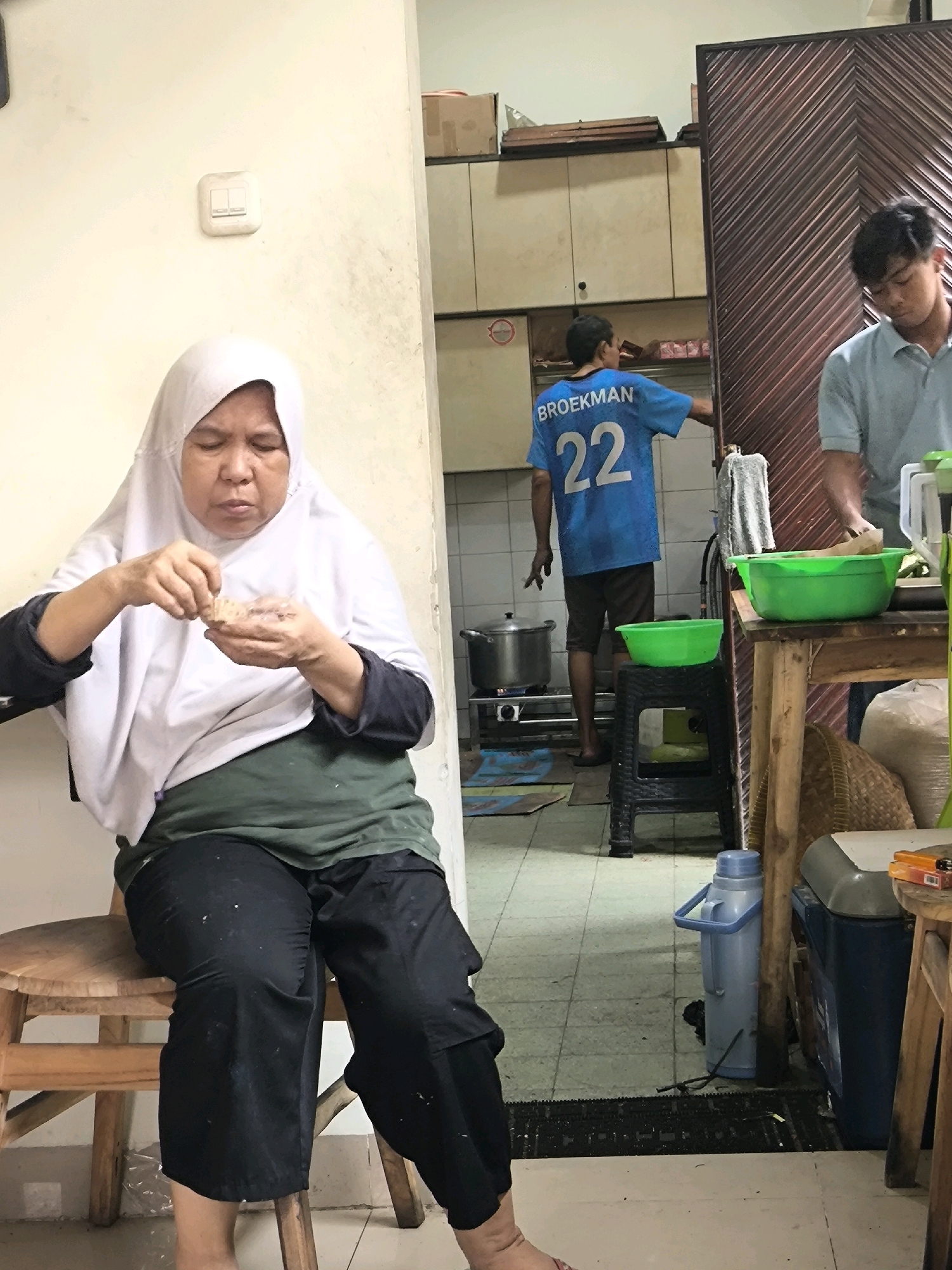 In a modest, brightly lit indoor space, possibly a kitchen or communal area in West Jakarta, Indonesia, an elderly woman wearing a white hijab and dark clothing is seated on a low wooden stool. She is holding a small piece of food in her hands and appears to be eating it, with her eyes cast downwards. To her right, in the background, a younger man in a light blue collared shirt stands at a wooden table, his hands engaged with something on the table, partially obscured. Further in the background, another man in a blue t-shirt with the number "22" and "BROEKMAN" printed on it is visible in a kitchen area. He is standing in front of a gas stove with a pot on it. The environment includes tiled flooring, cabinets, a green plastic bowl on the floor, a blue thermos, and various other household items scattered around. The lighting suggests it is daytime, with light coming from an unseen source, possibly a window or overhead lights.