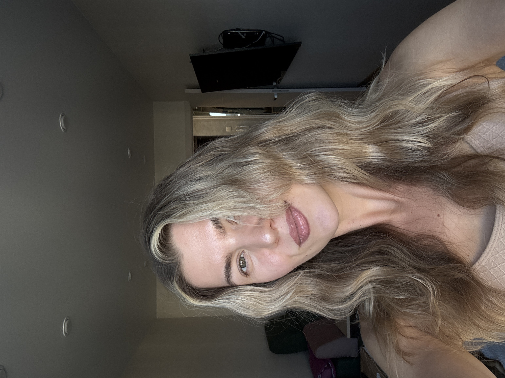 A young woman is the central subject of this indoor selfie, her head tilted slightly with a gentle, inviting smile. She has long, wavy hair, a mix of blonde and light brown highlights, cascading around her shoulders and partially obscuring the right side of her face. Her left eye, a light brown or hazel color, is visible and directly engaging with the camera. She has well-defined eyebrows and is wearing a glossy, nude-pink lipstick. Her skin appears clear and luminous. She is dressed in what looks like a light beige or pink top with a subtle, textured pattern on the fabric.
The background reveals a simple indoor setting. A plain, light gray wall is visible behind her, featuring several small, circular, silver-colored objects that could be decorative rings or light fixtures. In the upper portion of the frame, a black television or monitor is mounted on the ceiling, tilted downwards. A cluster of wires and cables is visible behind and around the mounted screen, suggesting electronic equipment. The lighting is diffused and appears to be artificial, casting soft shadows, hinting at an indoor environment possibly during the evening or with curtains drawn. No specific outdoor elements or visible text are present in the image.