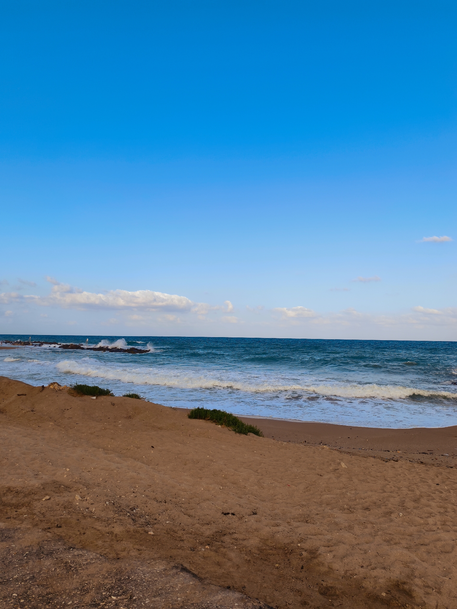The image shows a wide, sandy beach in Nahariyya, Israel, with the Mediterranean Sea in the background. The sky is a clear, bright blue with a few scattered white clouds. The sea is a deep turquoise, with gentle waves breaking on the shore and crashing against a rocky breakwater in the distance. There are no people visible in the image, and no specific objects or structures that would indicate the exact location within Nahariyya, other than it being a coastal area. The sand is a light brown, with some scattered pebbles and a few patches of sparse green vegetation growing on the dunes. The overall impression is one of a tranquil, natural landscape.