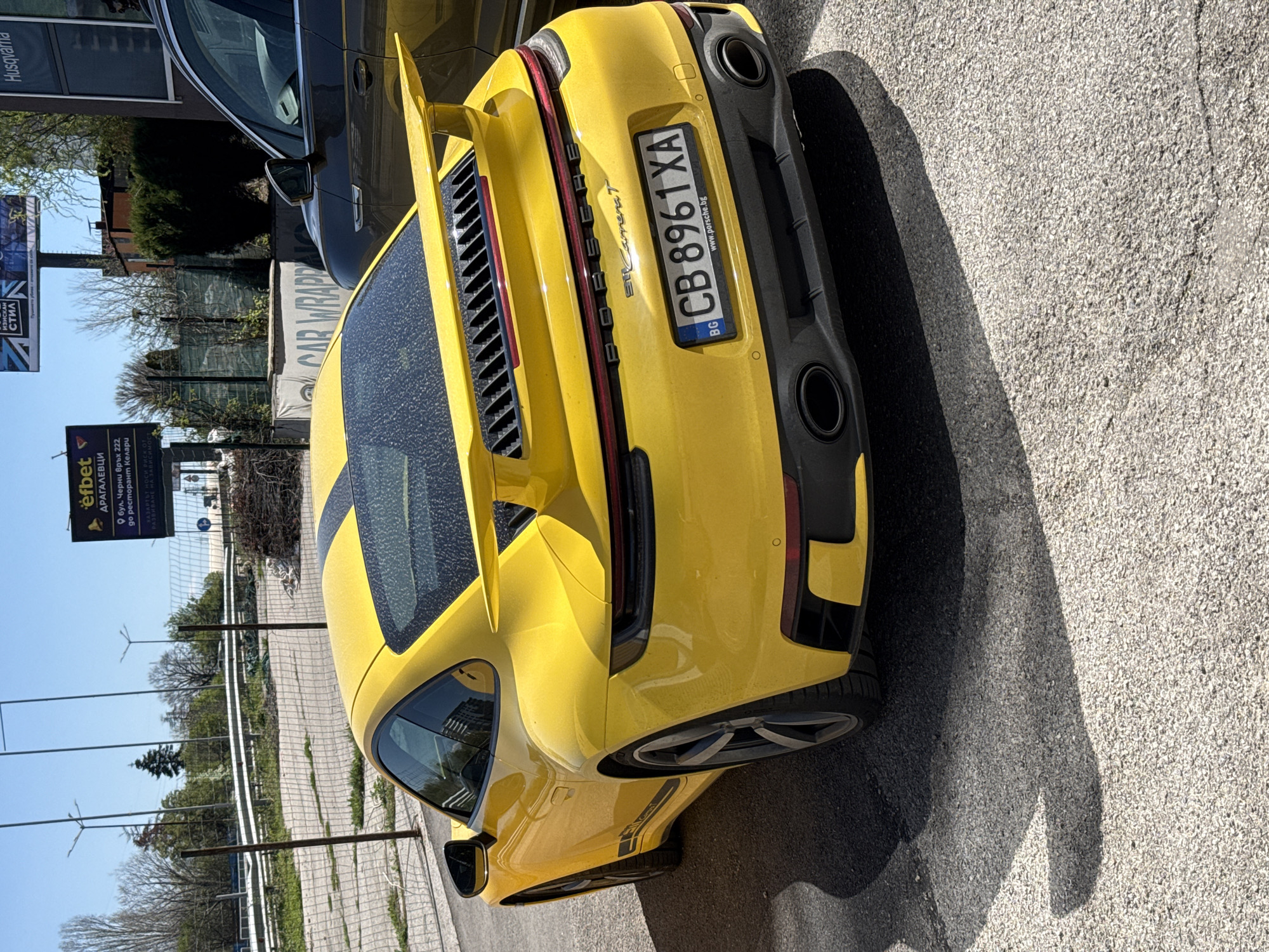 The image shows the rear of a yellow Porsche 911 Carrera T, parked on a paved surface. The Bulgarian license plate reads "CB 8961XA." The car is parked near a road with advertising signs for "Efbet" and "Husqvarna." The "Efbet" sign provides the location as "bul. Cherni vrah 222, do restaurant Kelari" in Sofia, Bulgaria. Other signs visible mention "CAR WRAP" and "IZISKAN Стил". A grey vehicle is parked next to the Porsche. The weather appears sunny, casting a shadow of the car on the road.