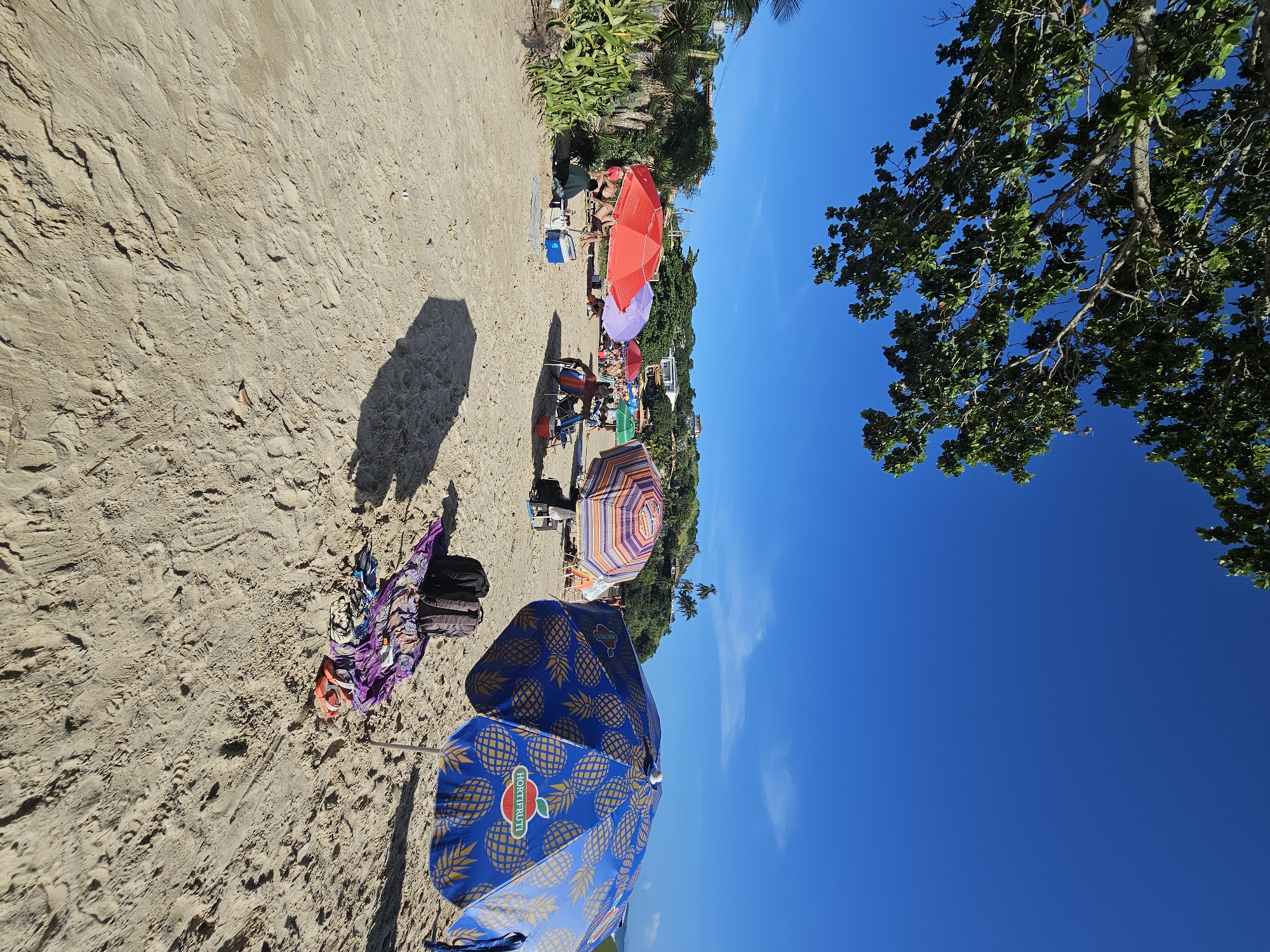 The image shows a sandy beach scene in Armação Dos Búzios, Brazil, with multiple beach umbrellas. The nearest umbrella has a blue pineapple design. A bag and clothing are on the sand near this umbrella. Further along the beach, there are other umbrellas in various colors like red, purple, and striped. People are relaxing under some of the umbrellas. Vegetation is seen in the background near some of the beachgoers, and there's a clear blue sky. Tree foliage is in the upper right corner.