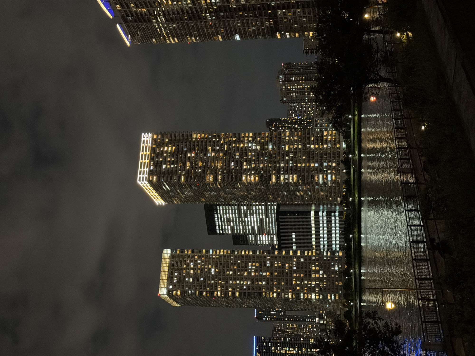 This is a nighttime photograph of a waterfront cityscape in Koto, Japan. The scene is dominated by numerous illuminated high-rise buildings, their windows glowing with warm yellow light against the dark sky. The buildings vary in height and architectural style, with some featuring sharp, modern designs and others appearing more rounded.
In the foreground, a dark, calm body of water reflects the lights from the buildings and the sky. A railing-lined walkway or promenade runs along the edge of the water, with scattered streetlights casting pools of illumination. Dark trees and foliage line the embankment, partially obscuring the lower portions of the buildings. The sky is overcast with dark clouds, contributing to the nighttime atmosphere. No people are visible in the frame.