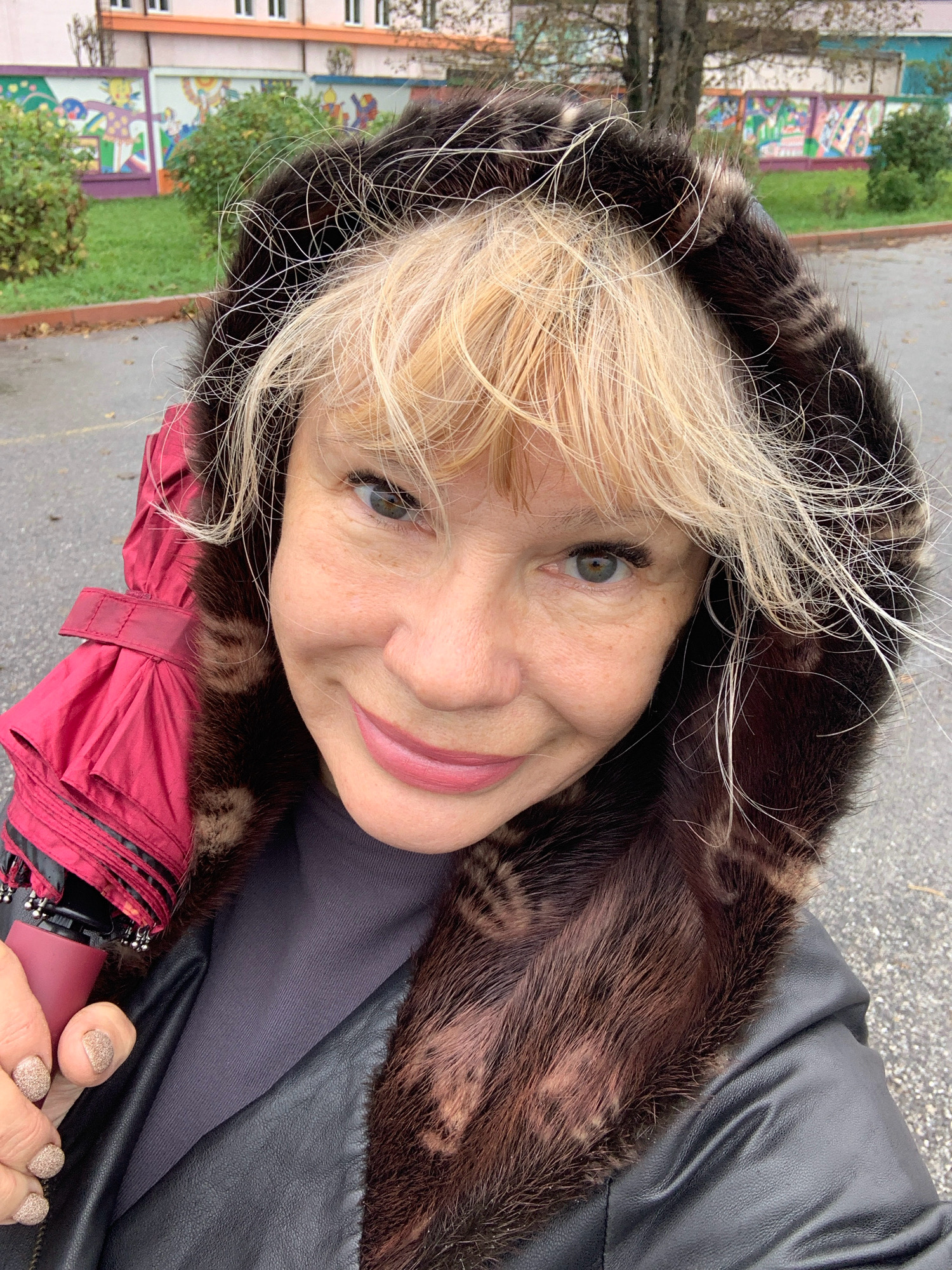 A close-up, selfie-style photograph captures a middle-aged Caucasian woman with blonde hair, wearing a black leather jacket and a hooded coat with a fur trim. She is holding a closed red umbrella in her left hand. The background shows a low building with windows and a colorful mural, accompanied by green bushes and a paved outdoor area. The lighting and foliage suggest an overcast day, possibly in autumn. The woman is looking directly at the camera with a slight smile. The scene is likely situated in a public area, perhaps near a school or community center given the mural.