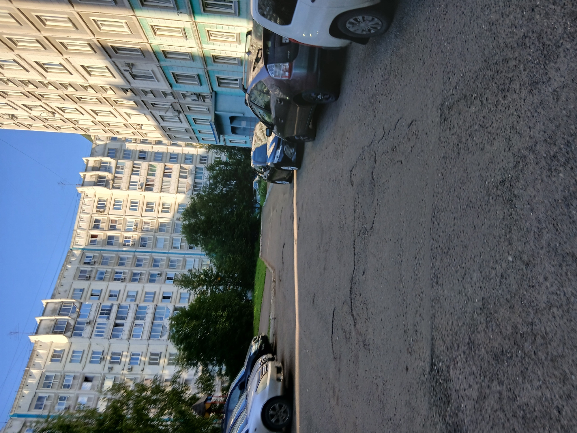 In a city setting, a view of residential buildings and parked cars is presented. The buildings, characterized by their light-colored facades and numerous windows, are multi-story structures. Some buildings exhibit a blue accent on their lower floors. In the foreground, a paved road surface is visible, showing signs of wear and tear, with cracks and uneven patches. Several cars are parked along the side of the road, including a silver sedan, a dark-colored sedan, and a dark-colored hatchback. Trees with green foliage are situated between the buildings and the road, adding a touch of nature to the urban landscape. The sky above is clear and blue, indicating fair weather conditions. The lighting suggests that it is daytime, with sunlight casting shadows.