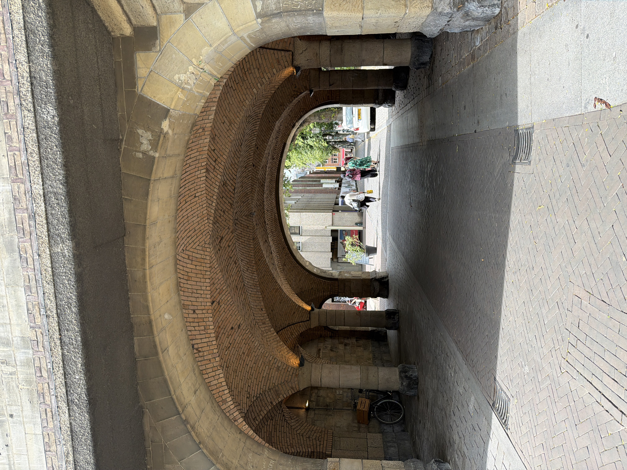 This is a photograph taken from beneath a brick-arched overpass, likely a bridge, on a sunny day in Roermond, Netherlands. The overpass is supported by thick stone pillars, with visible graffiti on some of them. The ground is paved with brick in a herringbone pattern, with a drain grate visible in the foreground. On the left side, under the overpass, a bicycle with a wooden crate on the back is parked next to a stone wall. The perspective looks out towards a street where a white car is parked on the right. A few people are walking away from the camera down the street, with buildings and trees visible in the background. The overall impression is of an urban environment with historical architectural elements.