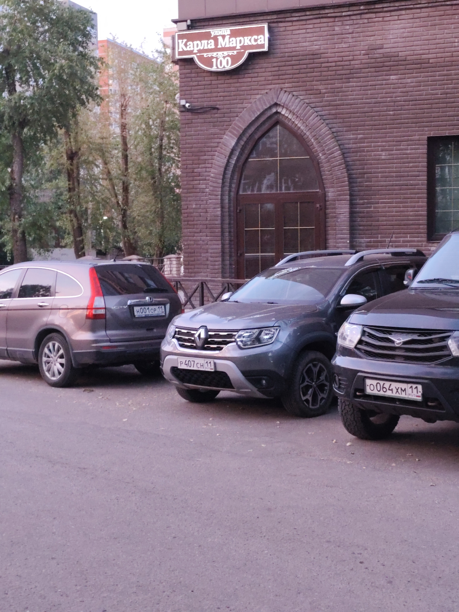 The image shows a street scene in Syktyvkar, Russia, with several cars parked on an asphalt road. In the background, a brick building with an arched entrance is visible. A street sign above the entrance reads "улица Карла Маркса 100," indicating that the location is Karl Marx Street, house number 100. There are no people visible in the image. The environment appears to be an urban setting.
