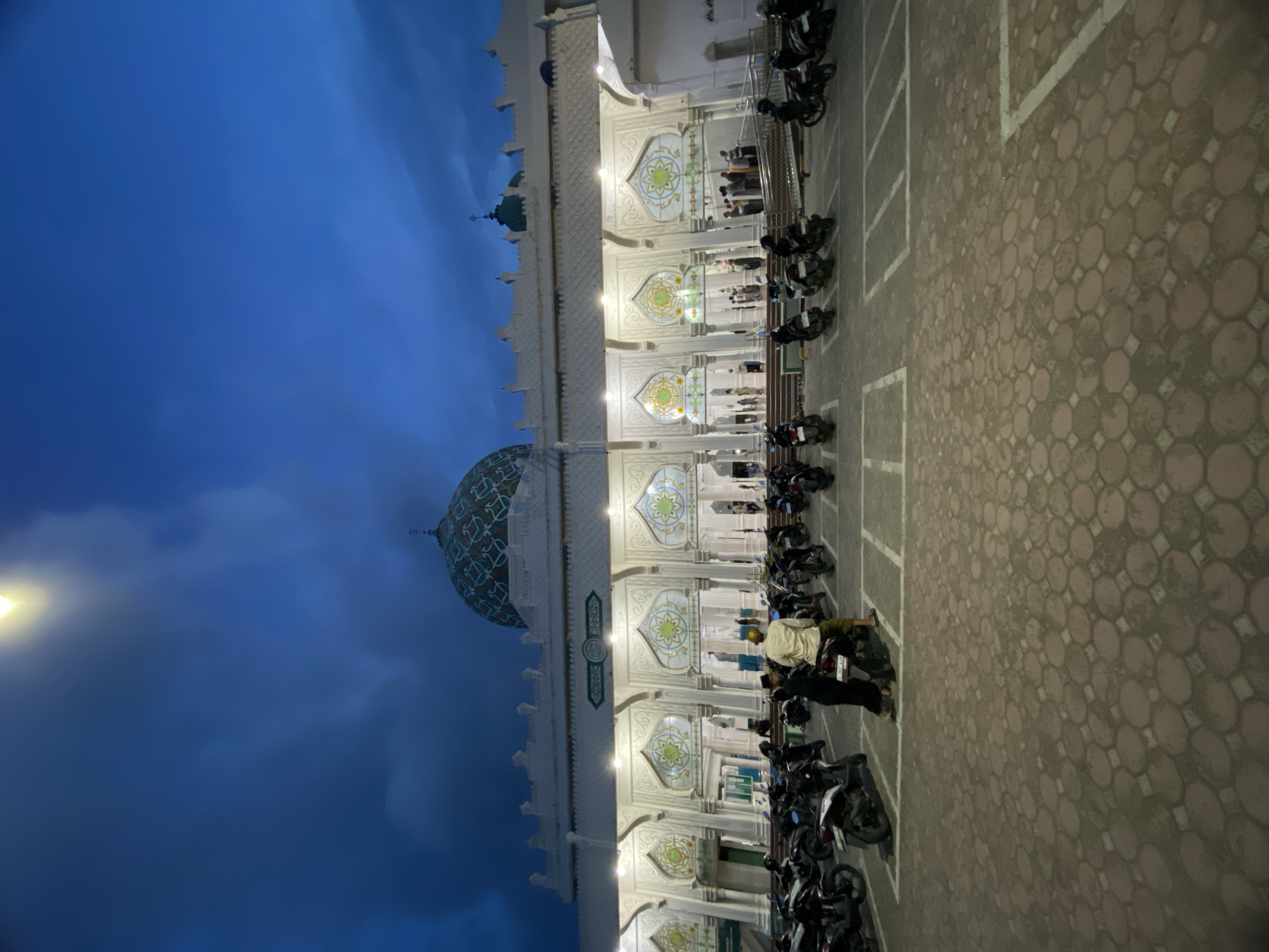 The image depicts a mosque in Lhokseumawe, Indonesia at dusk. A crowd of people are lined up to pray along the exterior of the mosque, in front of ornate windows. Motorcycles are parked in organized rows along the edge of a brick-paved lot. The sky is a deep blue.