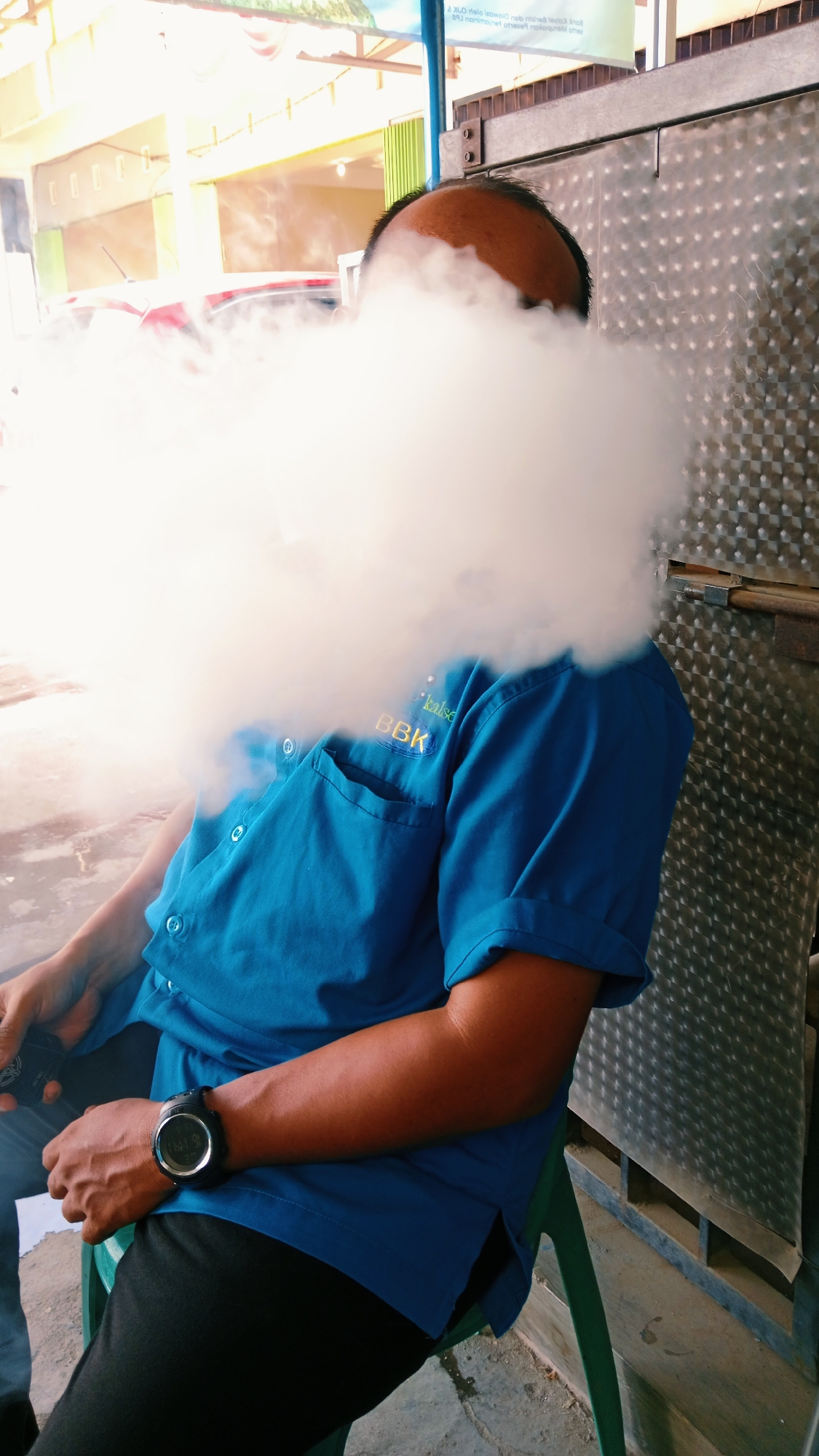 A medium shot shows a person sitting on a green chair, partially obscured by a large cloud of white vapor. The person wears a blue collared shirt with yellow text "BBK" and "kalsa" embroidered on the left breast. A black wristwatch with a circular face is visible on their left wrist. The person's head and upper body are covered by the vapor, but the forehead and scalp are visible through it. The background consists of a textured metallic wall to the right, a concrete floor below, and a blurred outdoor scene with buildings and a red vehicle in the distance to the left. The overall impression is that the person is likely at an outdoor establishment in Handil Bakti, Indonesia, possibly a cafe or shop, engaged in vaping or smoking.