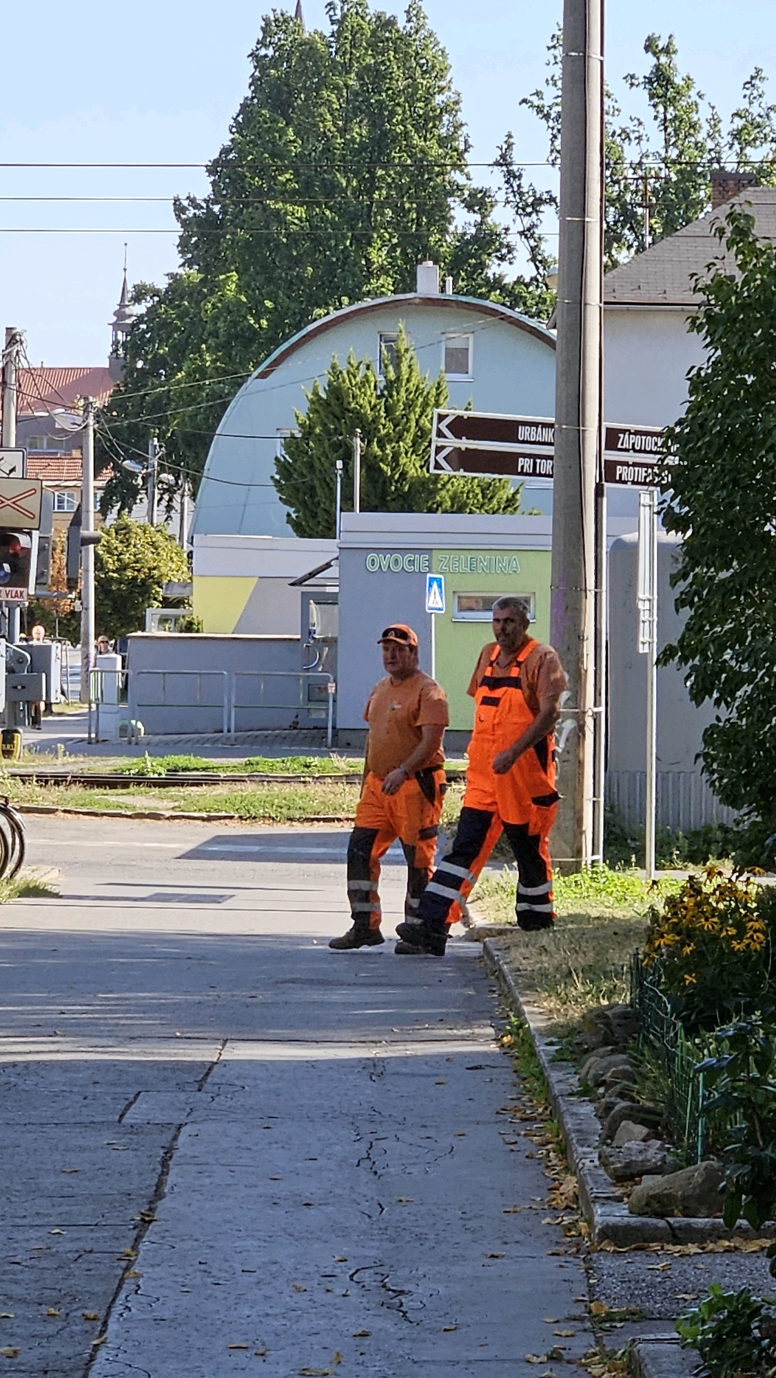 In Prešov, Slovakia, on a clear, sunny day, two male construction workers are captured mid-stride, walking from left to right across a paved road. Both are wearing high-visibility orange work uniforms, with the worker on the right in orange overalls and the worker on the left in orange trousers and a matching shirt. The worker on the right has a darker band around his pants near the ankle. They are walking past a curb lined with rocks and overgrown grass, with some yellow flowers visible in the background. Behind them, a light blue building with a curved roof is visible, marked with the text "OVOCIE ZELENINA," indicating it's a fruit and vegetable stand. A grey pole with multiple directional signs is also present near the building, with some signs appearing to point left. Further in the background, trees and buildings of the city are visible. The overall environment suggests an urban street or intersection.