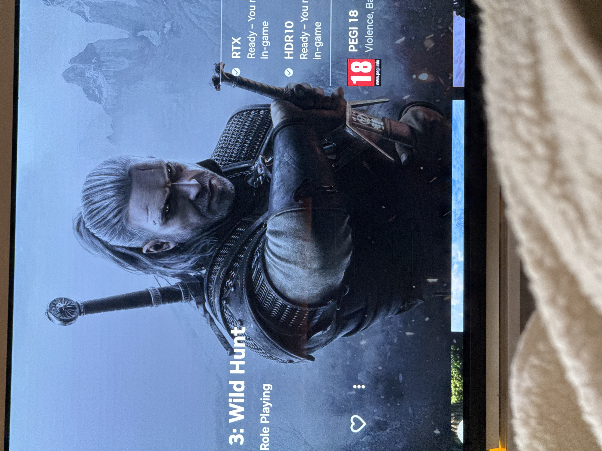 The image shows a TV screen displaying the game "The Witcher 3: Wild Hunt". The game's title and "Role Playing" genre are visible. The screen also indicates that RTX and HDR10 are ready in-game. A PEGI 18 rating for violence is shown. The main image is of Geralt of Rivia, a character from the game, holding a sword. No information on the city of Riga, Latvia is present.