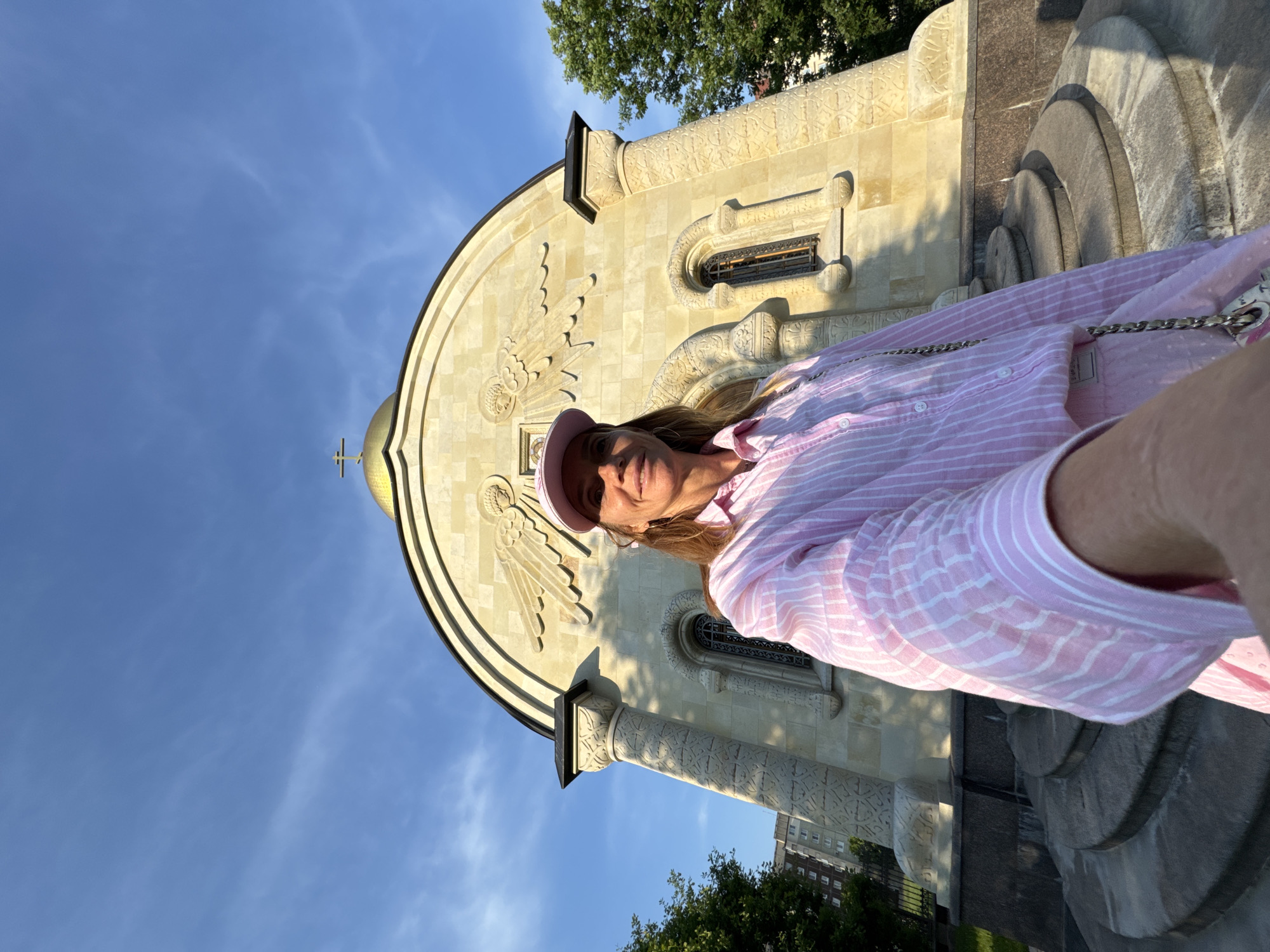 A woman stands in front of a golden-domed church in Kirishi, Russia, taking a selfie. She is wearing a pink and white striped button-down shirt and a pink baseball cap. The church is made of light-colored stone with intricate carvings and arched windows. The sky is blue with some wispy clouds, and trees are visible in the background. The woman is smiling and holding her phone in her right hand to take the picture. The perspective of the selfie is low-angle, with the church appearing large and imposing.