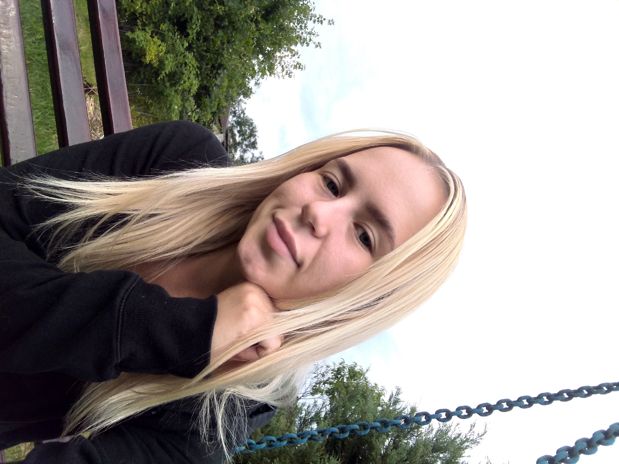 A young woman with blonde hair is sitting on a swing, outdoors. She is wearing a black hoodie and has her chin resting on her hand. In the background, there are trees and a cloudy sky. The swing's chains are visible in the upper right corner of the image. The location is Radisne, Ukraine.