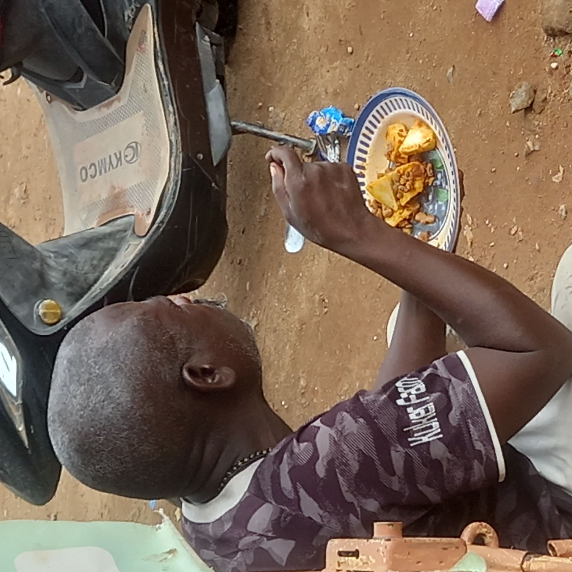 An overhead shot shows a man seated on the ground next to a Kymco scooter, eating from a plate of food with a spoon. The food appears to be yams and beans. The man is wearing a t-shirt with "KORE FSO" printed on the sleeve. The ground is a mixture of dirt and small stones. The picture was taken in Sardauna, Nigeria.