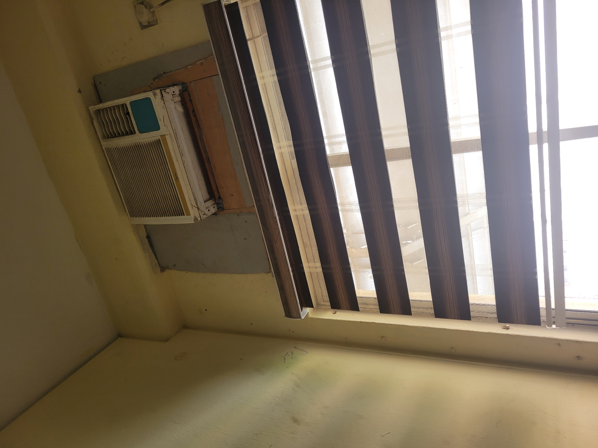 The image displays an interior room, likely in Abuja, Nigeria, during daylight hours. The main subjects are a window-mounted air conditioner unit and a window covered by vertical blinds.
The air conditioner, a white unit with a prominent gridded front and a turquoise-colored accent panel, appears aged and shows signs of wear and tear. A faint "Midea" brand name is visible on its lower right side. The installation around the unit is noticeably rough, with gaps filled by wooden planks and what looks like plaster or cardboard, suggesting a makeshift setup. Above the air conditioner, a power outlet or junction box is visible on the upper wall.
To the right of the air conditioner, a window is brightly illuminated by natural light, indicating it is daytime. The window is adorned with vertical blinds that feature a distinctive pattern of alternating dark brown striped sections and translucent beige or white sections, commonly known as zebra blinds. The blinds are partially open, allowing bright sunlight to flood into the room.
The room's walls are painted a light yellow or beige hue and exhibit significant signs of age and wear, including visible dirt, stains, and chipped paint. The floor, possibly concrete or painted, also appears worn with various marks. Some indistinct and illegible scribbles are present on the lower part of one wall. The overall impression is of a functional but somewhat older or poorly maintained space. No people or specific activities are visible in the scene.