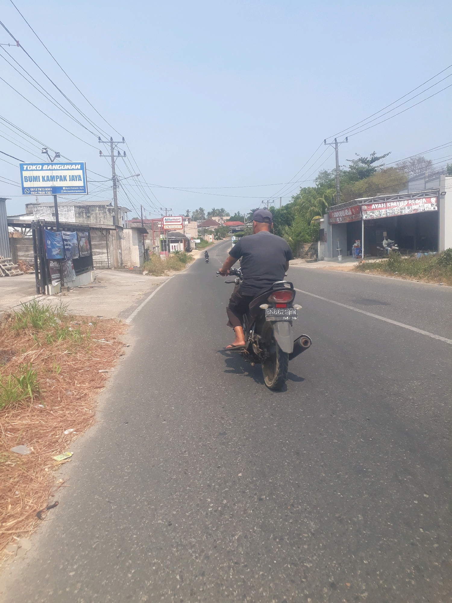 The image captures a sunny daytime scene on a paved road in Pangkalpinang, Indonesia. The perspective is from behind a person riding a motorcycle, looking forward. The rider, wearing a dark t-shirt, dark shorts, and a dark baseball cap, is operating a black and silver motorcycle with a visible license plate reading "BN 6629 HW". The motorcycle is in motion on the right side of the road.
To the left of the road, there is dry, straw-like grass and some scattered debris. Further left, a blue sign for "TOKO BANGUNAN BUMI KAMPAR JAYA" is visible, along with a metal gate and buildings. To the right of the road, there are several businesses. One sign reads "AYAM KREMES PRAGA". Another sign displays "Alfamart". Behind these businesses, trees and houses are visible. Utility poles with overhead wires line both sides of the road. The sky is clear and pale blue, suggesting a bright day. The overall environment appears to be a commercial and residential area along a main road.