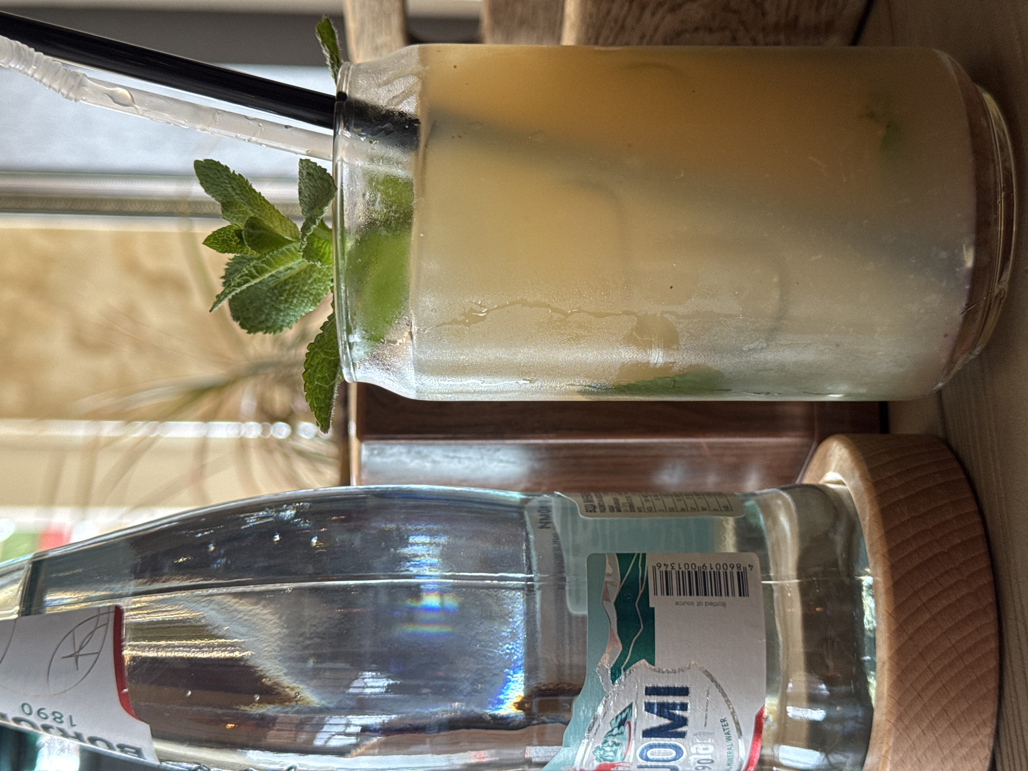 A close-up, eye-level shot presents a drink and a water bottle, oriented horizontally. The drink, likely a cocktail or mixed beverage, is contained in a clear glass with a metal straw, suggesting it is served cold, possibly on ice. Mint leaves garnish the glass. Positioned below the drink is a clear glass water bottle with a light blue and white label. A wooden coaster sits at the bottom of the water bottle. The scene appears to be set indoors, possibly at a table in a cafe or restaurant, based on the blurry background elements. The location is noted as the city of Sirius, Russia. No people are visible.