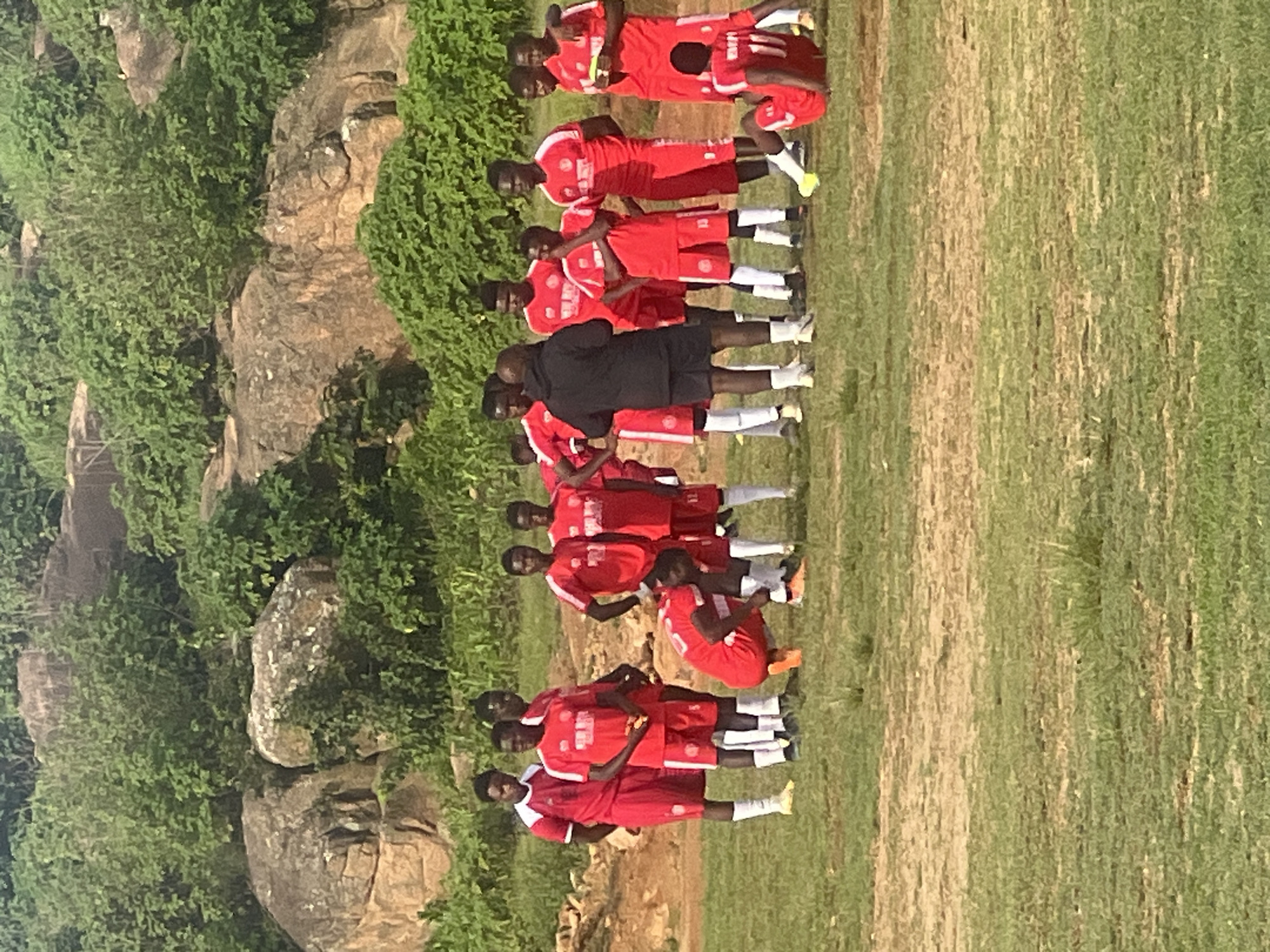 The image depicts a group of young men in matching red football kits gathered on a grassy field. The field appears to be a local training ground, possibly in Jos, Nigeria, with a rocky, vegetated hillside in the background. Some of the men are standing, while others are in various poses, suggesting a training session or a break. One individual, dressed in a black hoodie and shorts, appears to be addressing the group. The overall atmosphere is casual and focused on the sport.