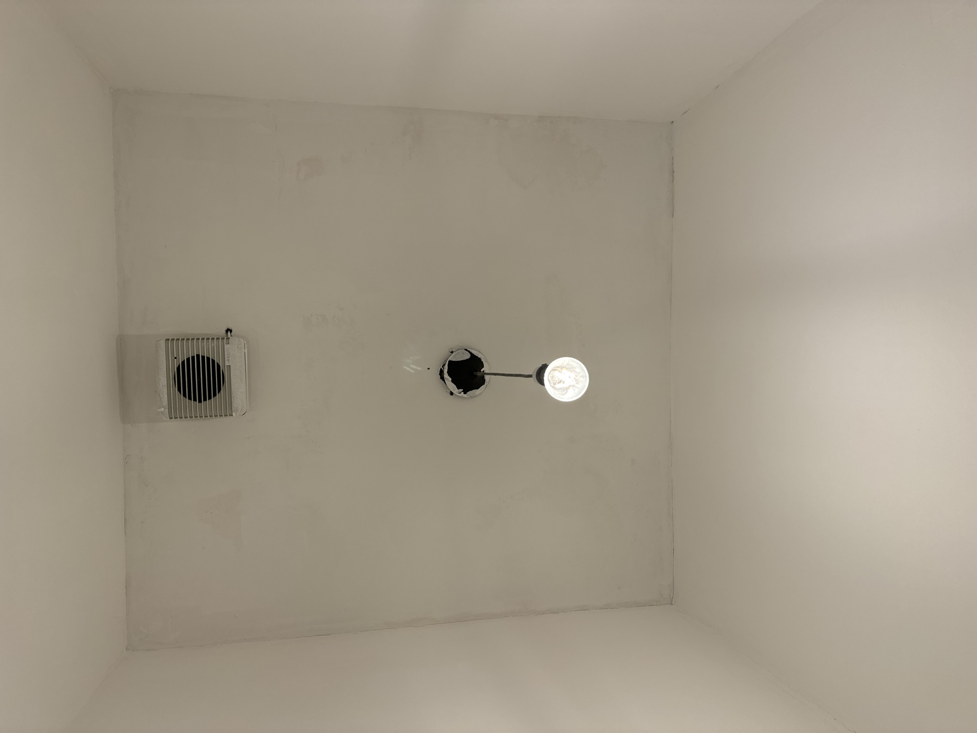A lightbulb hangs from a wire suspended from a hole in a plain white ceiling. Above and to the left of the lightbulb is a rectangular ventilation grille. The ceiling appears slightly textured with some discolored patches, possibly water stains, and there are visible lines where the ceiling meets the walls. The walls are also plain white. The scene is indoors, likely in a room with unfinished or minimally finished surfaces. There is no indication of the specific location within the city of Waterford, Ireland.