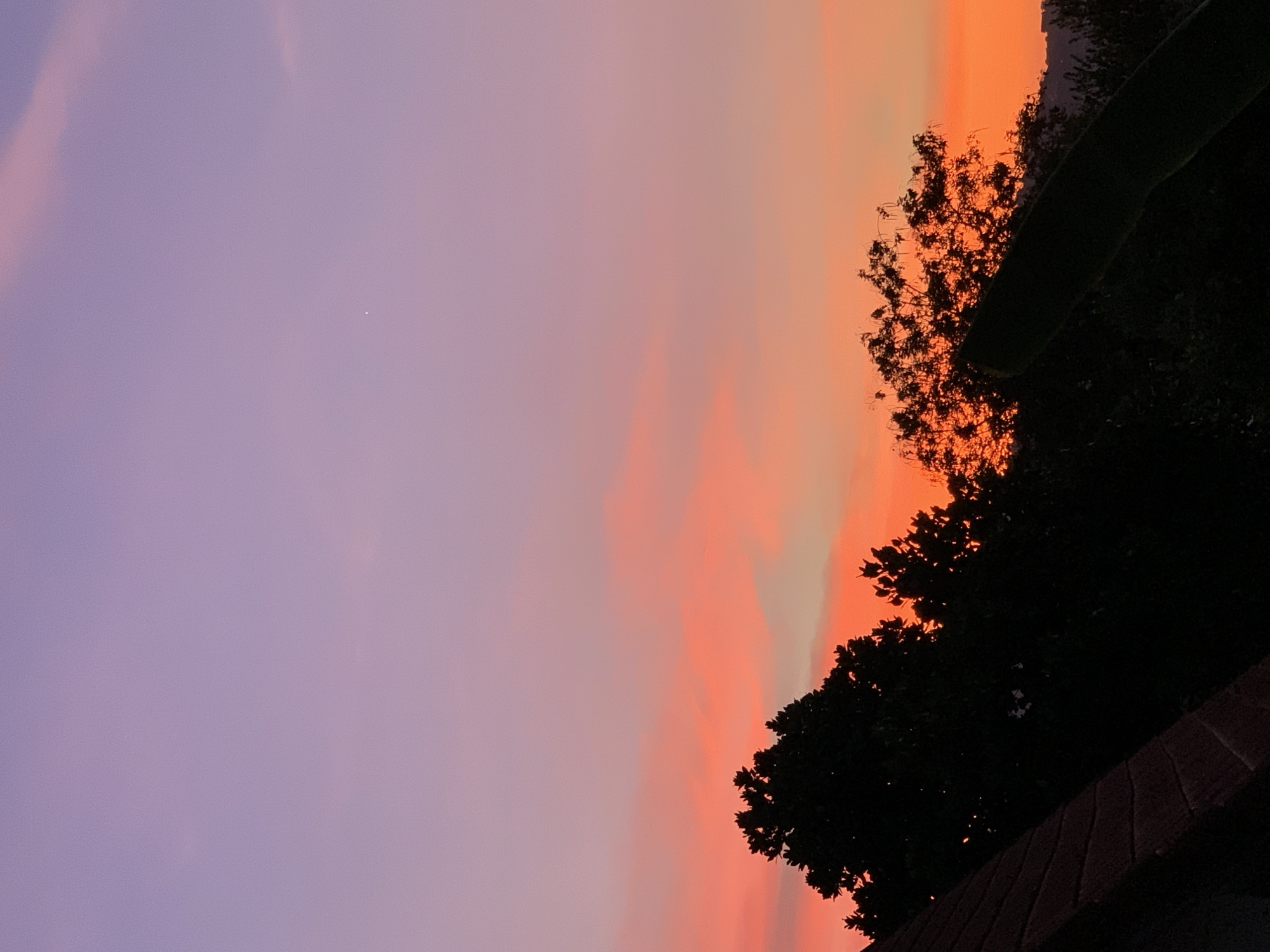 The image shows a view of the sky at sunset in บ้านบางรักษ์, Thailand. The sky transitions from purple on the left to orange on the right, with streaks of orange and pink in the center. The silhouette of trees occupies the right side of the image, partially obscuring the view of the sky. A small, bright spot is visible in the purple portion of the sky, potentially a star or planet. A portion of a dark, tiled surface is visible in the bottom right corner.