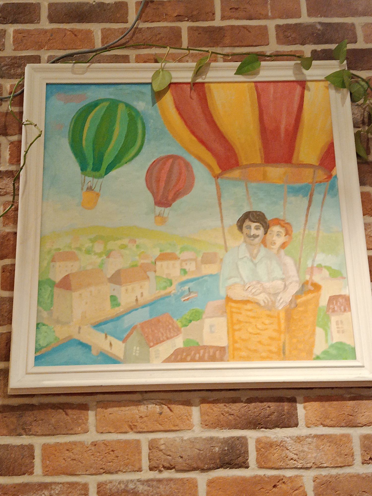 The image shows a framed painting hanging on a brick wall. A vine with green leaves drapes across the top of the frame.
The painting depicts a man and a woman in the basket of a hot air balloon, which is striped in yellow and red. Two other hot air balloons are in the background, one green and one red. Below the balloons is a cityscape with a river running through it. The buildings are pastel colors.
There is no information given that suggests this picture came from Moscow, Russia.