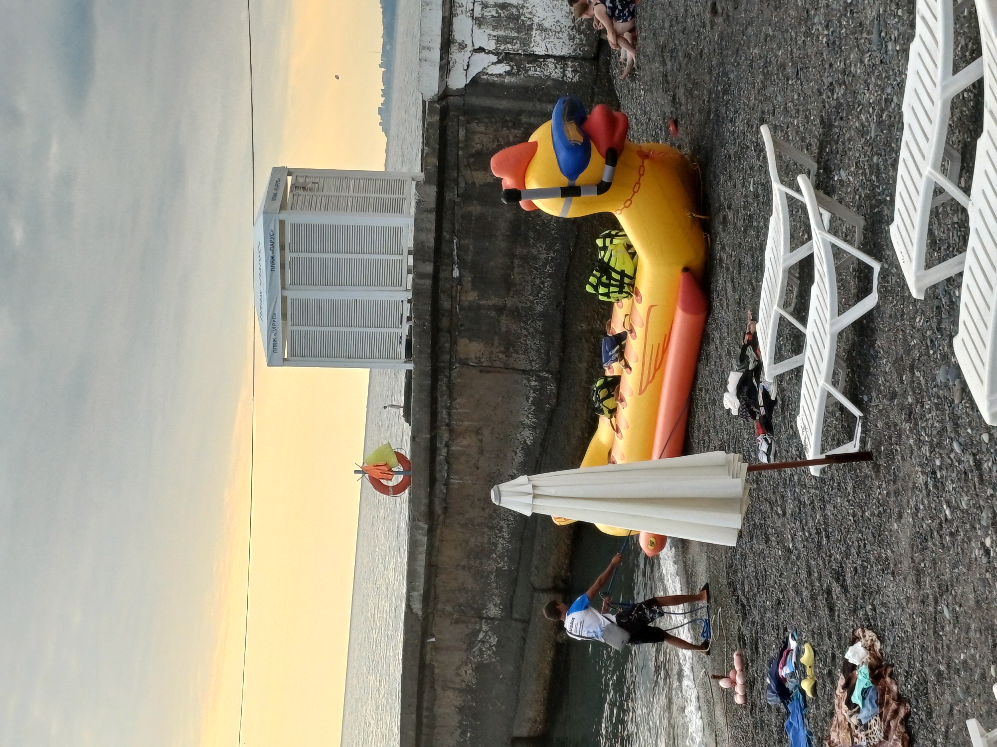 This is a landscape photo taken at a beach in Sochi, Russia, likely during sunset due to the warm glow in the sky. The scene is set on a pebble beach with a concrete embankment separating the beach from the sea.
The main subjects in the foreground are a large, inflatable yellow duck toy wearing a snorkel mask and goggles, and a closed white beach umbrella. A man, possibly an attendant or rescuer, is seen near the umbrella, holding a rope. There are several pieces of clothing and other beach items scattered on the ground. Two white lounge chairs are visible further up the beach, and a white changing booth or small structure with louvered doors stands near the water's edge. In the background, the sea stretches out to the horizon, with a distant city skyline visible.
The activity appears to be a quiet evening at the beach, with preparations for closing or just winding down. The overall mood is calm and peaceful.
Notable details include the soft, warm light of what appears to be sunset, indicating the time of day. The setting is clearly a beach, with the sea, embankment, and beach furniture. The inflatable duck toy suggests a family-friendly environment or a water park. The text visible on the changing booth, "ПЛЯЖ «ПАРУС»," translates to "BEACH 'SAIL'."