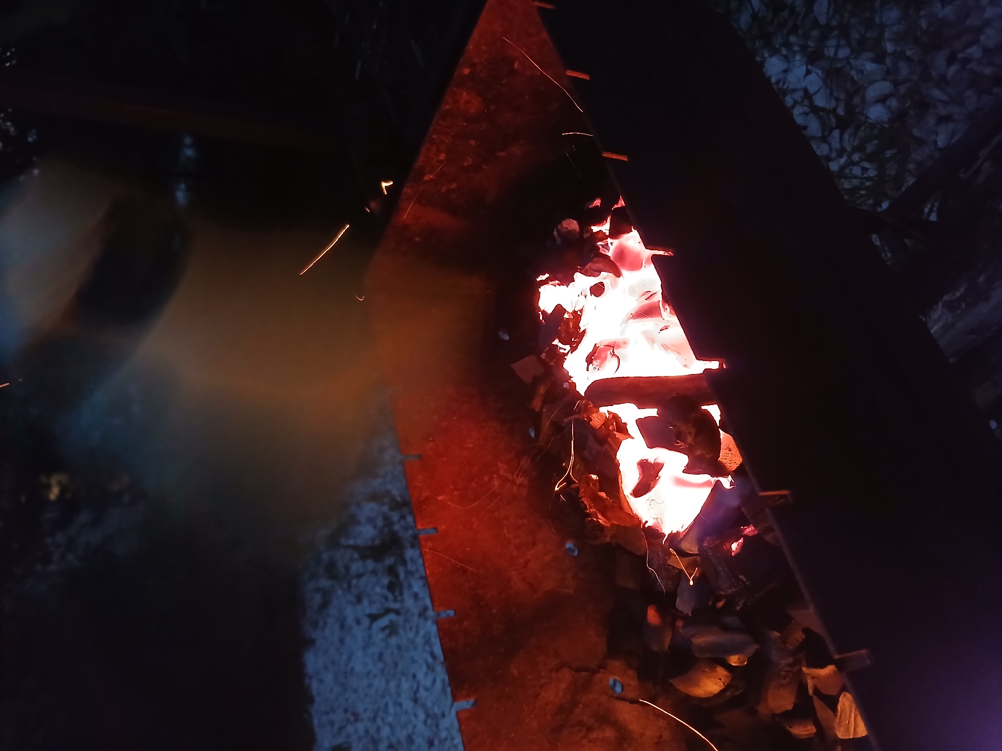 The image captures a close-up view of a fire burning in what appears to be a metal fire pit or brazier. The primary subject is the intense glow of the burning embers and flames, which radiate a bright white and orange light, illuminating the surrounding charcoal and wood fragments. Sparks are visible, arcing away from the main fire, suggesting a lively and active flame. The fire pit itself is a dark, metallic structure, with a triangular opening through which the fire is viewed. The edges of the metal are dark and textured, with visible rivets or perforations along one side.
To the left of the fire pit, there is a dark, reflective surface, possibly water, with a subtle orange glow reflecting on it, likely from the fire. Further to the left, the scene transitions to a dark, blurry background with hints of a lighter, hazy area. To the right and behind the fire pit, the background is dark and textured, with a pattern that suggests gravel or small stones, interspersed with dark shapes that could be foliage or shadows.
The overall impression is of a nighttime scene, indicated by the darkness of the surroundings and the prominence of the artificial light from the fire. The image conveys a sense of warmth and the primal energy of a bonfire. There are no people visible in the image, and no text is present. The setting appears to be outdoors, given the gravelly texture and natural darkness. The context of Verkhneye Dubrovo, Russia, suggests a rural or semi-rural outdoor environment.