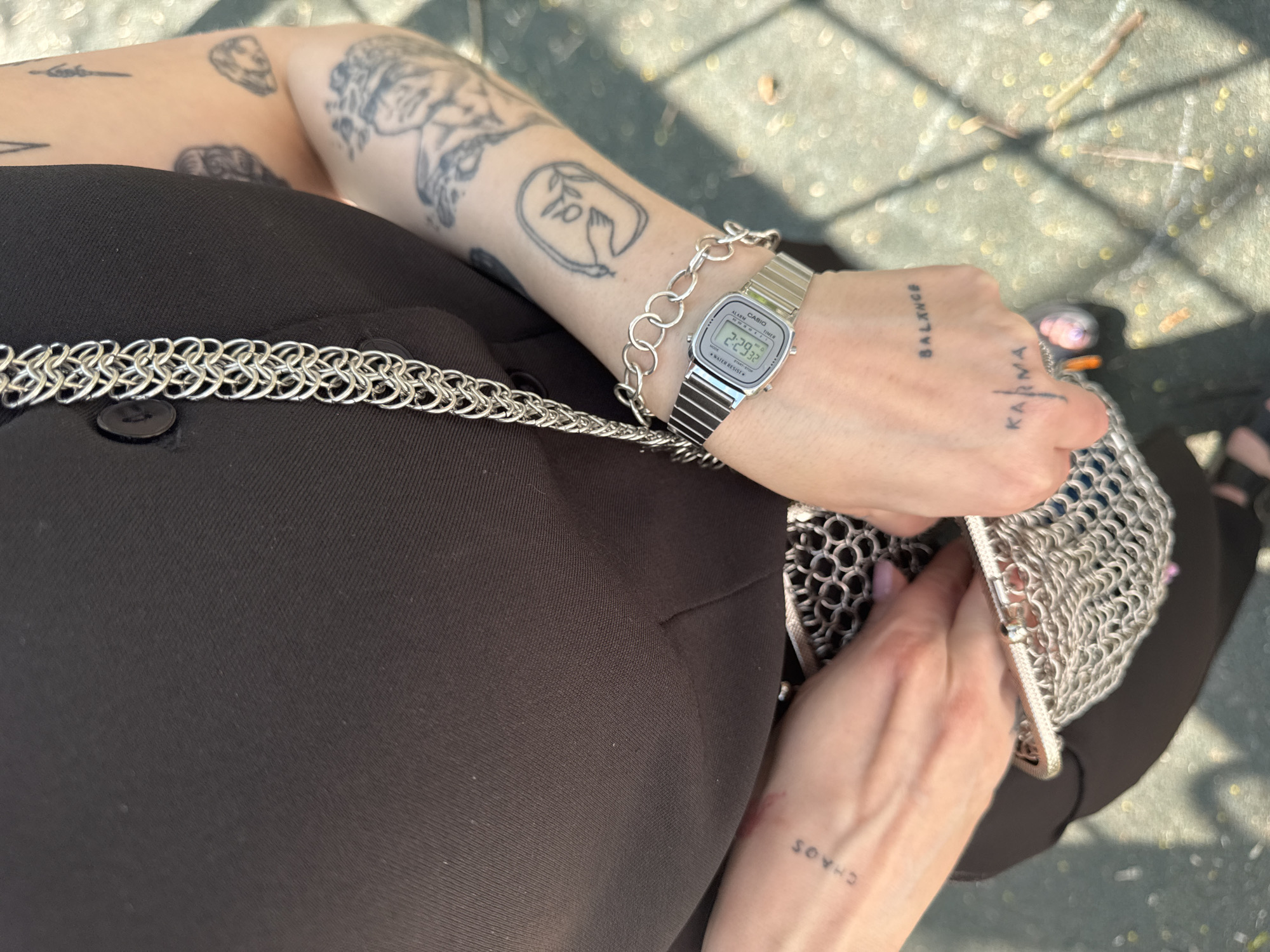 A person wearing a black blazer is holding a silver, chainmail-style purse. The person has several tattoos on their arms, including text that reads "BALANCE," "KARMA," and "CHAOS." They are also wearing a silver digital watch and a silver chain bracelet. The location appears to be outdoors, possibly on a paved surface. The time on the watch reads "2:29:32." It's not possible to determine if the location is in Moscow, Russia from the given image.