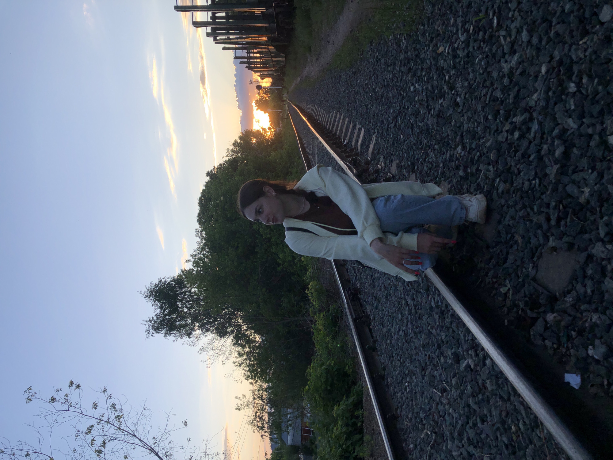 The image, taken in Chaikovsky, Russia, captures a young woman sitting beside a train track during sunset. She is positioned on the gravel ballast next to one of the steel rails, leaning against it with her body angled towards the viewer. She is dressed in light blue jeans, a dark top, a cream-colored jacket or cardigan with brown trim, and white sneakers. Her right hand, featuring red nail polish and a ring, rests near the track. Her expression is calm and neutral.
The train tracks, composed of metal rails and wooden ties, recede into the distance, where they meet a vibrant orange and yellow sunset. The track bed is covered with dark gray gravel. Dense green trees and foliage flank both sides of the tracks, with a particularly prominent stretch of trees on the left side. In the far distance, partially obscured by the trees and silhouetted against the bright horizon, are faint structures that resemble train cars or industrial buildings. A few small, distant houses are also visible through the trees on the right.
The sky transitions from a soft blue overhead to warm hues of orange and yellow near the horizon, characteristic of dusk or late evening. A few wispy clouds are visible, reflecting the sunset colors. The scene conveys a tranquil, natural setting at the end of the day. There is no visible text in the image.