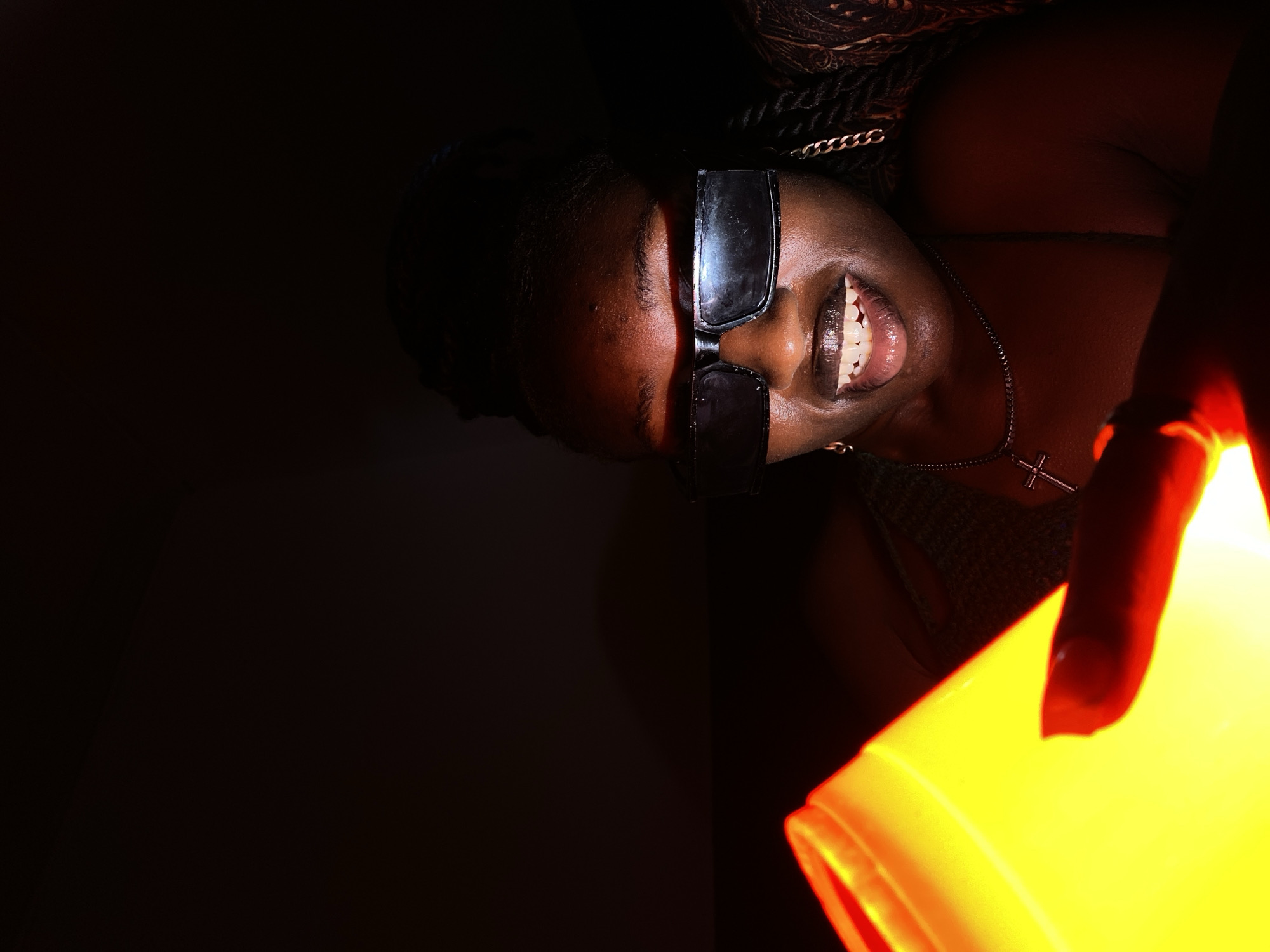 A close-up, medium shot captures a Black woman in sunglasses illuminated by a bright yellow light source in a dimly lit environment. The woman's mouth is open, revealing her teeth, and her eyes are hidden behind dark rectangular sunglasses. She is wearing a necklace with a cross pendant and a chain with a distinct link pattern, possibly an earring, is visible. A portion of a patterned fabric is visible on her right side. The light source, a cylindrical object glowing intensely yellow and orange, occupies the left side of the frame and casts a strong red hue on the woman's skin. The background is dark and indistinct, suggesting an indoor setting at night, potentially in Bar Jirgi Summa, Nigeria.