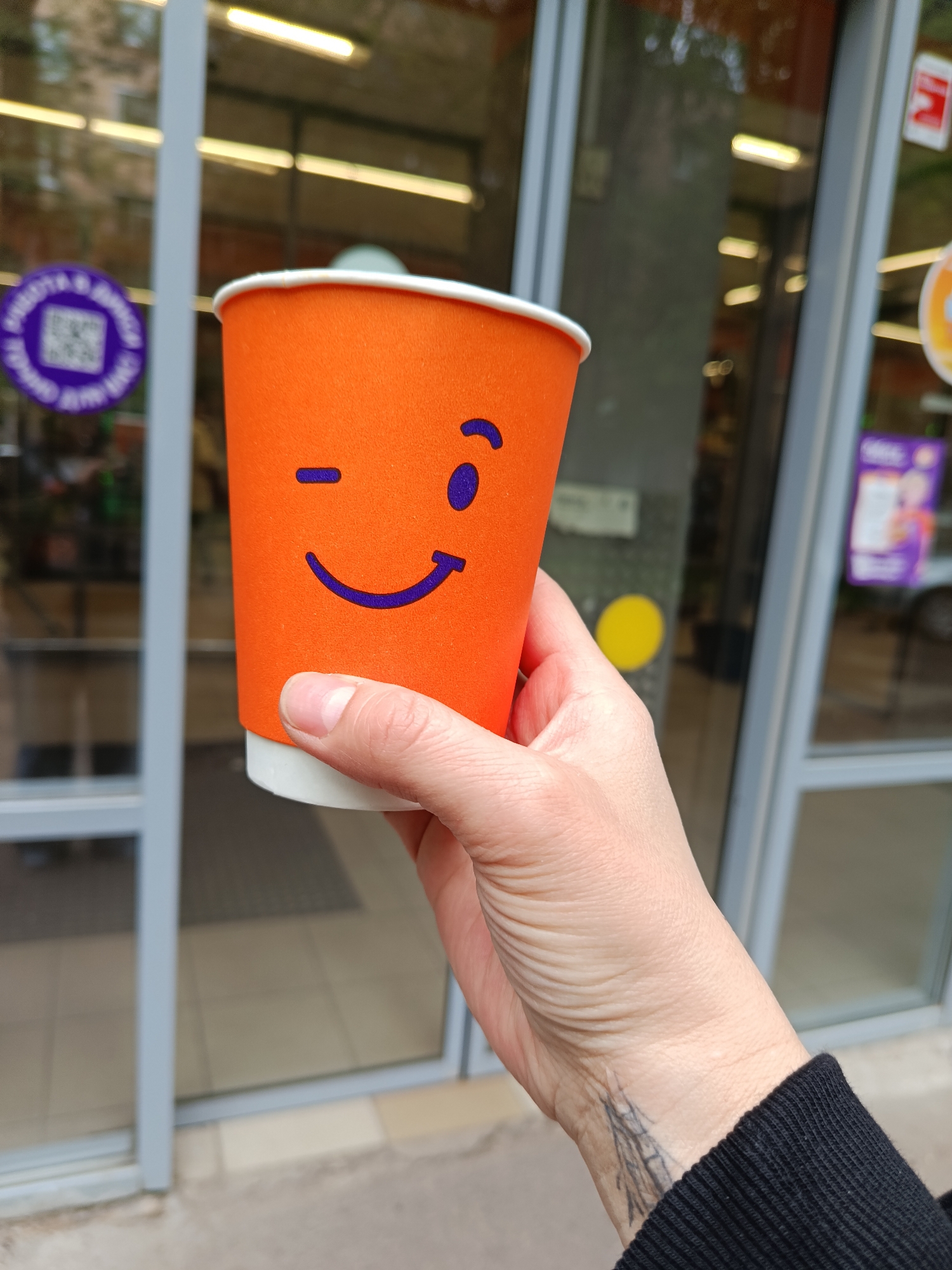 A person's hand holds an orange paper cup with a playful winking smiley face design against a blurred background. The cup features a blue winking eye and a blue smiling mouth on its vibrant orange surface. The hand, wearing a black sleeve, grips the lower part of the cup, revealing a dark, abstract tattoo on the wrist.
The setting appears to be outdoors, in front of a commercial building with large glass doors or windows. The glass reflects bright horizontal interior lights and hints of an indoor space with a doormat and tiled floor. On the left glass pane, a circular purple sticker with a QR code and some indecipherable text is affixed. Another blurred sign, partially orange and purple, is visible on the right glass pane, along with a yellow circular sticker on a central pane. The environment suggests a daytime scene, possibly overcast, in a city. The provided location context is Moscow, Russia.