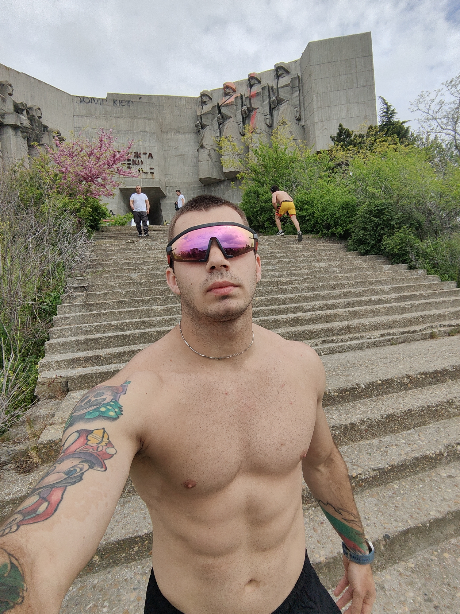 A shirtless man stands on the bottom steps of a long staircase leading to a concrete monument in Varna, Bulgaria. The monument, in the background, features relief sculptures and graffiti. Several people ascend or descend the stairs, including a man in yellow shorts. The foreground man wears sunglasses and a necklace and displays tattoos on both arms. Lush greenery surrounds the staircase, including a blooming pink tree. The sky is overcast.