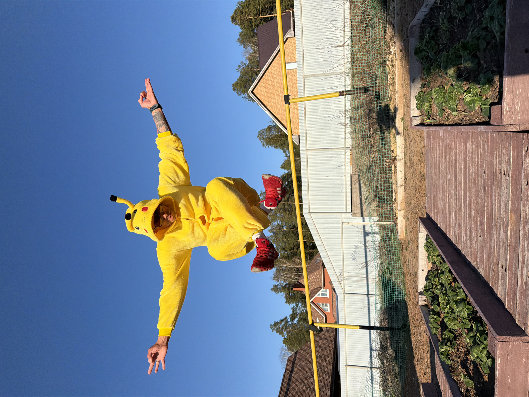 A man dressed in a Pikachu costume is captured mid-air, seemingly jumping, against a clear blue sky. He is wearing red sneakers and the Pikachu costume includes a hood with Pikachu's features. His left arm is raised with his fingers forming a hand gesture while the other arm is extended out to the side. A tattoo is visible on his raised arm.
The background shows a residential area. There is a house with white siding and a brown roof, and a yellow-colored pole structure runs vertically and horizontally. Behind the house, trees are visible. To the right, there is a wooden structure filled with green plants. The location is Chaikovsky, Russia.