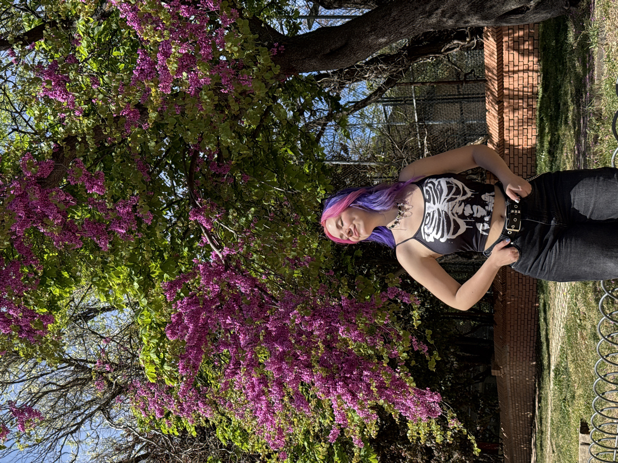 A young woman with dyed pink and purple hair, wearing a black tank top with a white skeleton print and black jeans with a studded belt, stands in front of a large tree with vibrant purple flowers. The background includes a brick wall, a metal fence, and a grassy area. The presence of redbud trees in bloom suggests that the image was taken in the spring. While the scene evokes a park-like setting, it is not possible to confirm the specific location in Madrid, Spain, solely from the image.