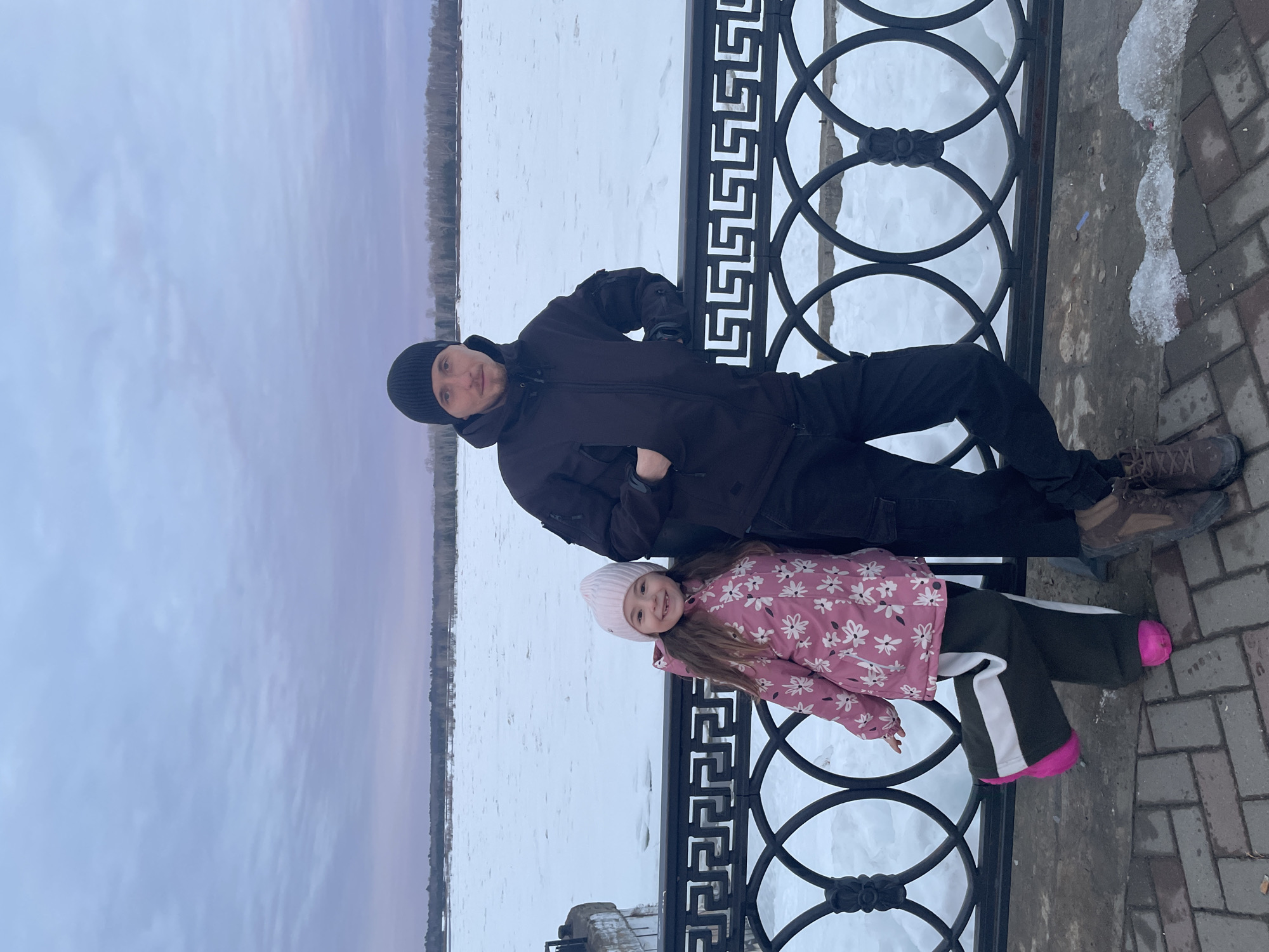 The image depicts an adult male and a young girl standing on the Sarapul Embankment, overlooking the frozen Kama River in Sarapul, Russia. The man, appearing to be in his late 20s to 30s, wears a black beanie, a dark hooded winter jacket, dark cargo pants, and dark brown lace-up boots. He is leaning his left arm on a black metal railing, with his right hand in his jacket pocket, and stands with his left leg slightly bent. He gazes directly at the camera with a neutral expression.
To his left stands a young girl, approximately 4-7 years old, with long brown hair. She wears a light pink knitted beanie, a pink winter jacket adorned with a white floral pattern, dark green pants with a prominent white stripe along the outer seam, and bright pink boots or shoes. She is smiling broadly at the camera, revealing a gap in her front teeth, and stands with her right leg lifted and bent at the knee.
The black metal railing, separating the subjects from the river, features a distinctive design: the upper horizontal bar is decorated with a repeating Greek key (meander) pattern, while the lower section consists of interwoven circular motifs. Behind the railing, the wide Kama River is completely frozen, covered in a vast, uneven expanse of snow and ice. A treeline of bare deciduous trees marks the distant riverbank on the horizon. The sky above is overcast, displaying gradients of pale blue and soft purple. The ground beneath the subjects is a paved walkway composed of reddish-brown and gray bricks laid in a herringbone pattern, with visible patches of melting snow and ice. The overall environment indicates cold, winter conditions.