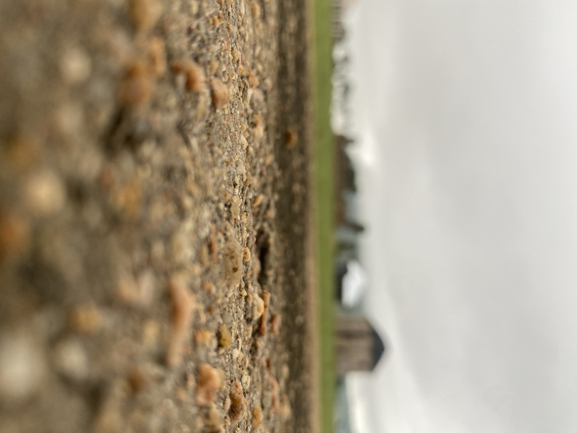 The image is a close-up, low-angle shot of a gravel surface in the foreground. The gravel is composed of small, multi-colored stones, predominantly in shades of brown, tan, and white. The background is out of focus, showing a blurry green field and several out-of-focus buildings, one of which appears to be a barn with a dark roof. The sky above is overcast and appears grey or white, suggesting a cloudy day. There are no people or animals visible in the image. The scene depicts an outdoor environment, possibly a rural or semi-rural setting. This image does not contain any information that could identify its location in Jalingo, Nigeria.