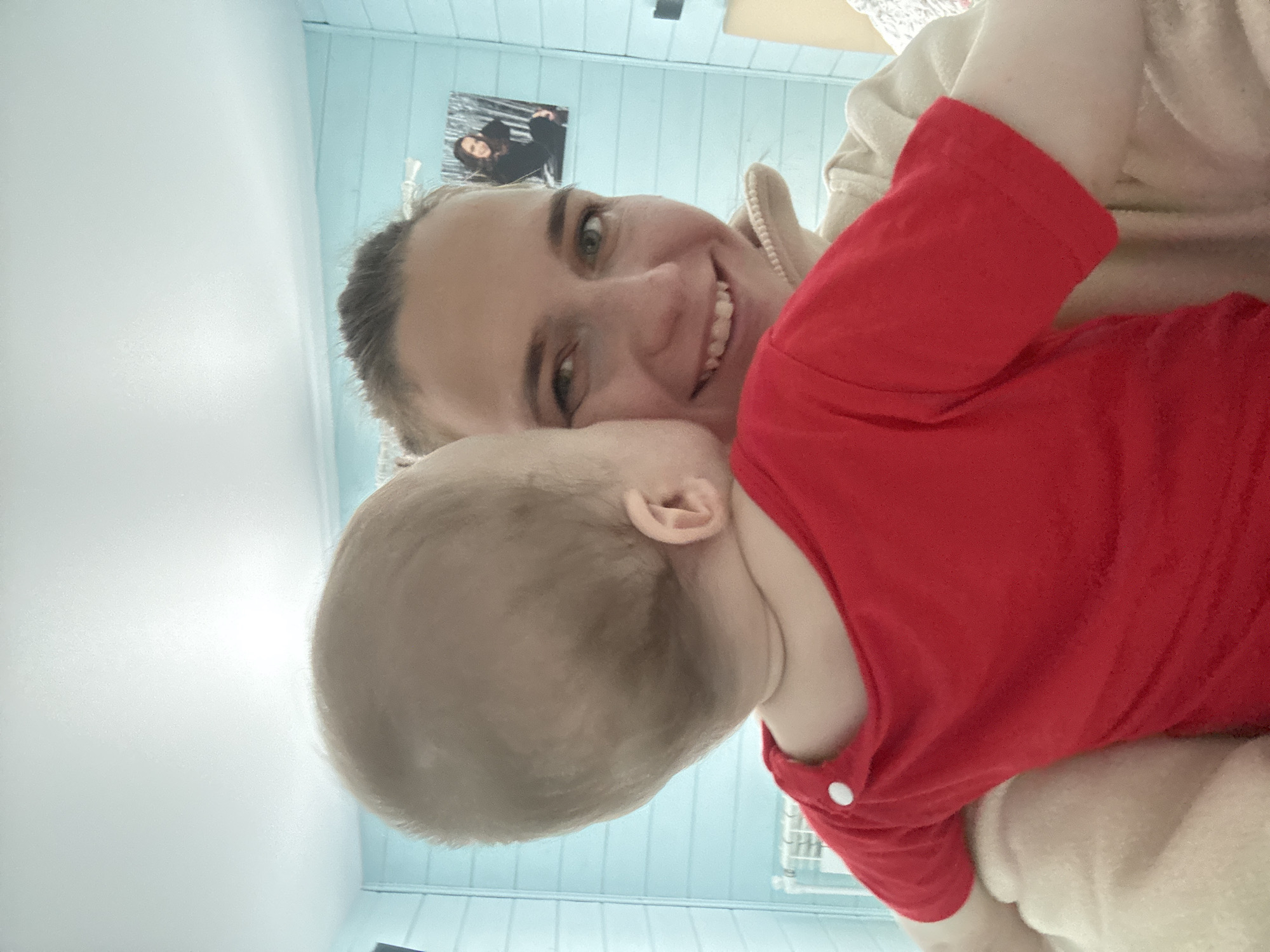 A woman with fair skin and dark blonde hair pulled back in a ponytail is smiling while holding a baby. The baby has a light complexion, blonde hair, and is wearing a red shirt. The woman is wearing a tan-colored sweater. The background consists of light blue paneled walls, a white ceiling, a framed picture on the wall behind the woman, and a white radiator. The location is possibly an interior space in Раздолье, Russia.
