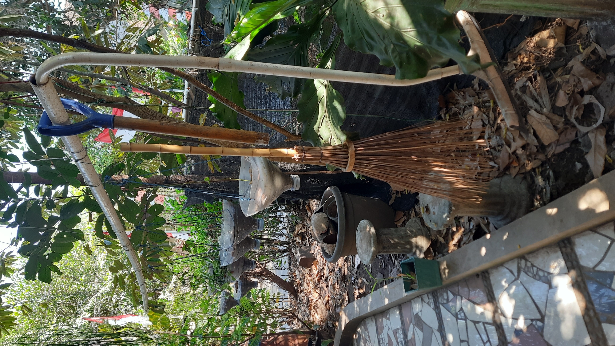 The image depicts an outdoor scene in Sumberarum, Indonesia, likely a yard or garden area. In the foreground, a tiled pathway runs along the left side of the frame, with a concrete planter box containing a small green plant. Further back, a large dark brown pot is filled with rocks and coconut husks. To the right of the pot, a broom made of dried bamboo stalks leans against a tree trunk. Above, a horizontal pipe structure, possibly for supporting hanging plants, extends across the image. Several repurposed plastic bottles, cut and inverted to serve as planters, hang from this structure. A shovel with a blue handle is propped against the pipe. The ground is covered in dry leaves and debris. In the background, lush green foliage and trees are visible, with hints of red and white Indonesian flags fluttering in the distance. The overall impression is of a somewhat unkempt but functional outdoor space, possibly a home garden.