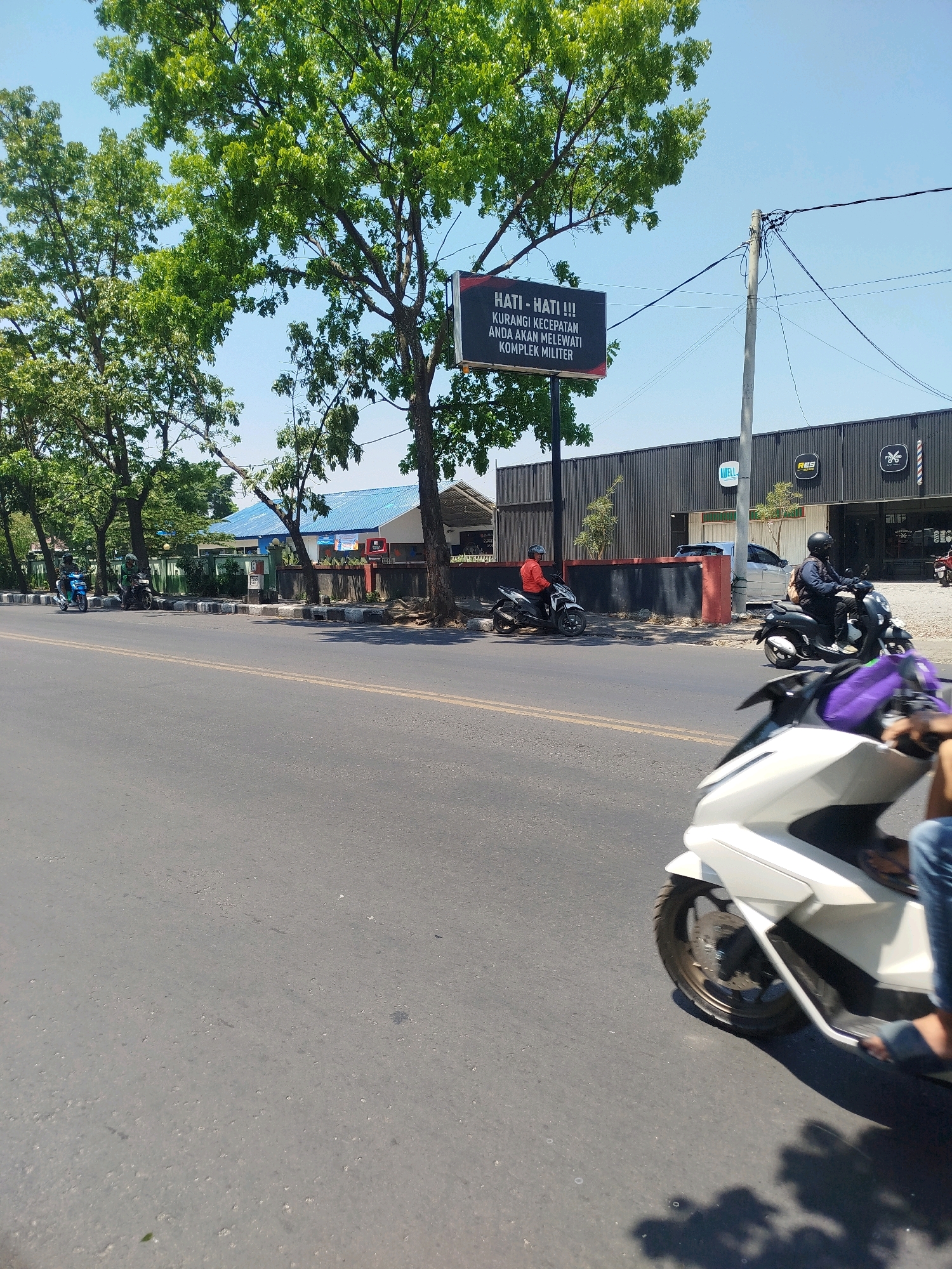 This is a photograph taken on a sunny day, likely in the city of Bandung, Indonesia. The image captures a street scene with several motorcycles, buildings, trees, and a clear blue sky. In the foreground, a white scooter is partially visible, with its front wheel and part of the rider's legs and feet in view. The rider is wearing blue jeans and sandals.
Further down the road, another motorcycle with a rider wearing a dark jacket and helmet is seen. To the left, a motorcycle in orange and white is parked. Beyond that, two more motorcycles are on the side of the road, with riders wearing helmets.
On the right side of the street, there is a modern-looking building with a dark facade and several signage. One sign is for "WELL," another for "RES," and a third with a barber pole logo. The building appears to be a commercial establishment.
On the left side of the road, there are several trees providing shade. Behind the trees, a building with a blue roof and white walls is visible. A prominent black billboard is suspended between trees and a utility pole, displaying text in red and white that reads "HATI - HATI !!! KURANGI KECEPATAN ANDA AKAN MELEWATI KOMPLEK MILITER" which translates to "BE CAREFUL !!! REDUCE YOUR SPEED YOU WILL PASS THROUGH MILITARY COMPLEX."
The road itself is asphalt, marked with yellow lines. The environment suggests a typical urban or suburban setting in Indonesia, with a mix of commercial buildings, residential areas, and lush greenery. The presence of the military complex warning sign indicates proximity to a military installation.