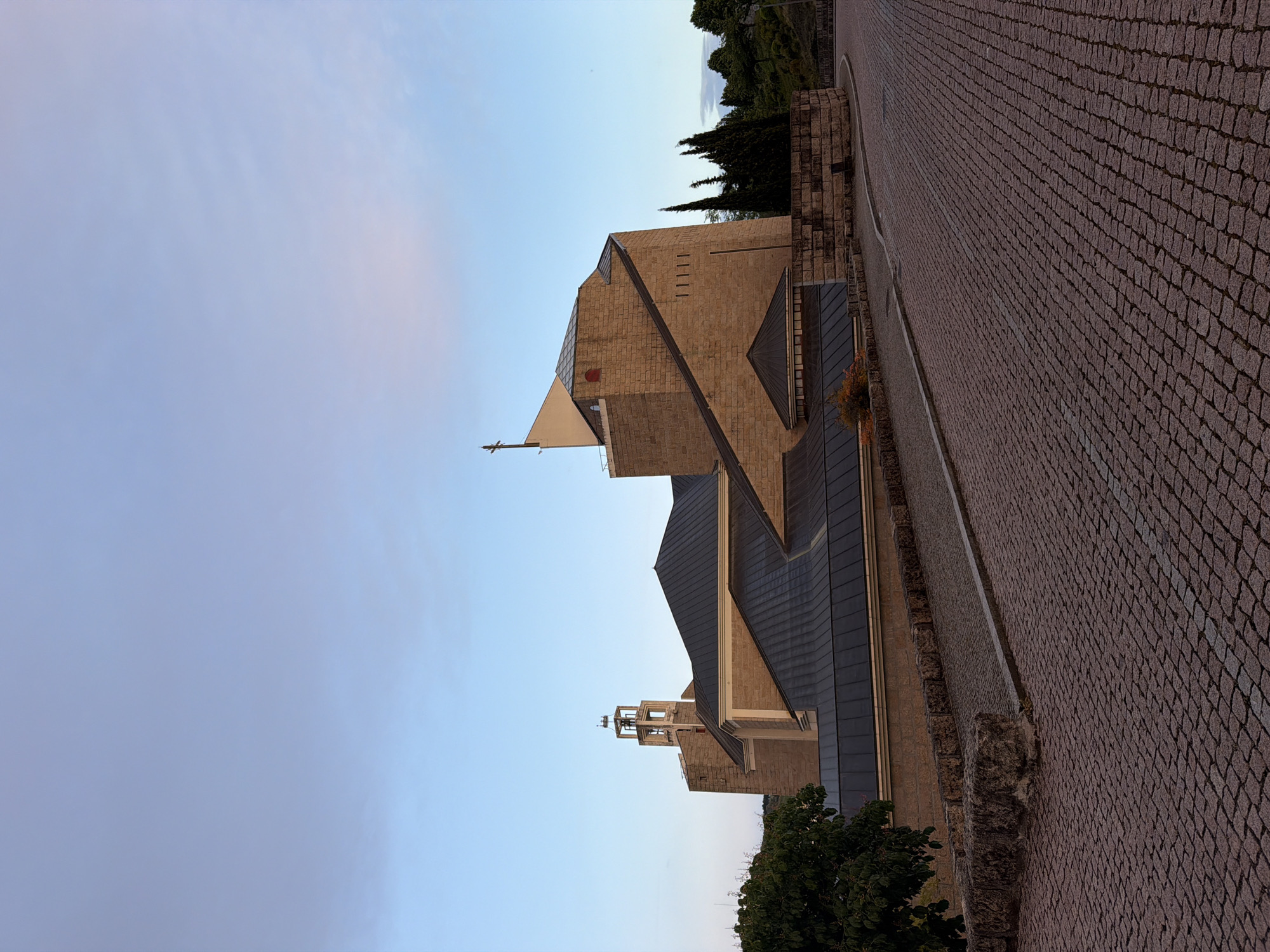 The image shows the Igreja de Santa Maria, a church in Bragança, Portugal. The building is constructed from tan-colored stone blocks with multiple sloping roofs covered in dark gray tiles. A cross is at the top of the main structure. A bell tower is visible near the bottom of the image. In the foreground is a cobblestone street with gray granite blocks. To the right of the church, a tiered stone wall sits adjacent to the street. To the left of the church is a pale blue sky. Vegetation, including trees, is visible in the lower left and upper right of the frame. The picture is taken from a low angle.