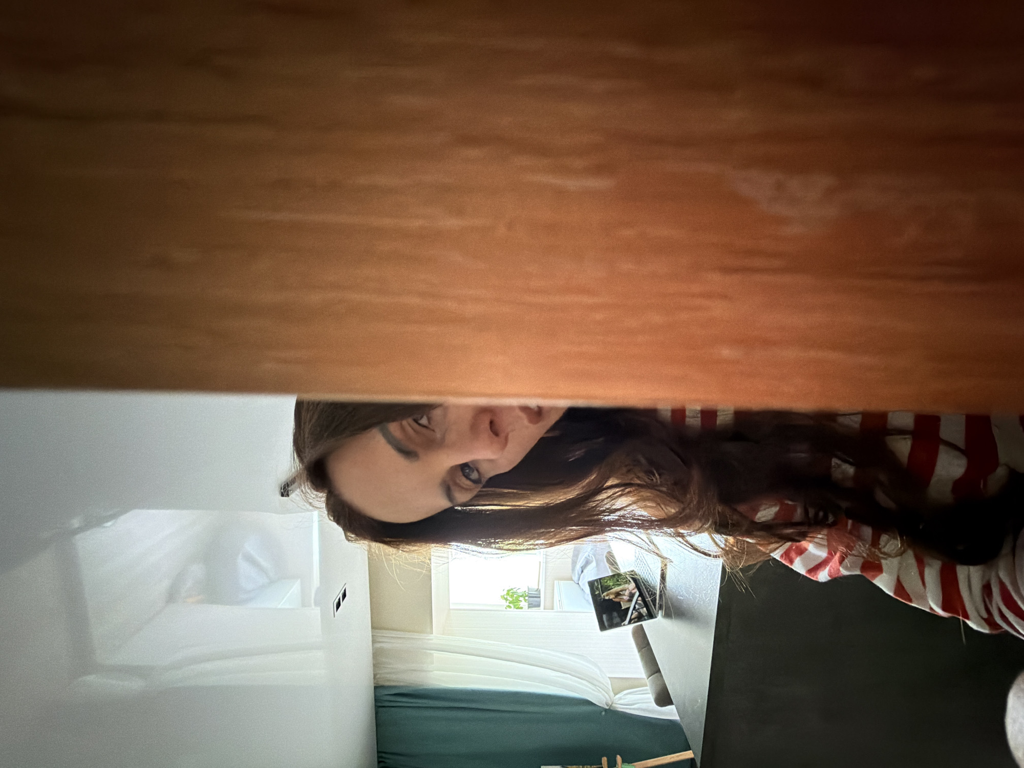 An adult female, positioned on the left side of a prominent reddish-brown wooden vertical structure (likely a doorframe), occupies the left-center of the frame. Her head is angled, with her visible left eye, nose, and part of her mouth directed towards the viewer. She has long, dark brown wavy hair and wears a long-sleeved garment with horizontal red and white stripes. The wooden structure occupies approximately 30-40% of the right side of the image.
In the background, a brightly lit interior room is visible. A large window, adorned with white sheer curtains and a teal-colored curtain pulled back on the left, allows natural light to enter. A small potted plant with green foliage rests on the windowsill. In the mid-ground, a dark-colored, rectangular table or counter holds an open silver laptop. The laptop screen displays an image featuring what appears to be two women, one with blonde hair, in an outdoor setting. A grey upholstered chair is partially visible behind the table. The ceiling above is white, reflective, and features two recessed rectangular light fixtures. The overall scene depicts a casual interior environment in the city of Birobidzhan, Russia.