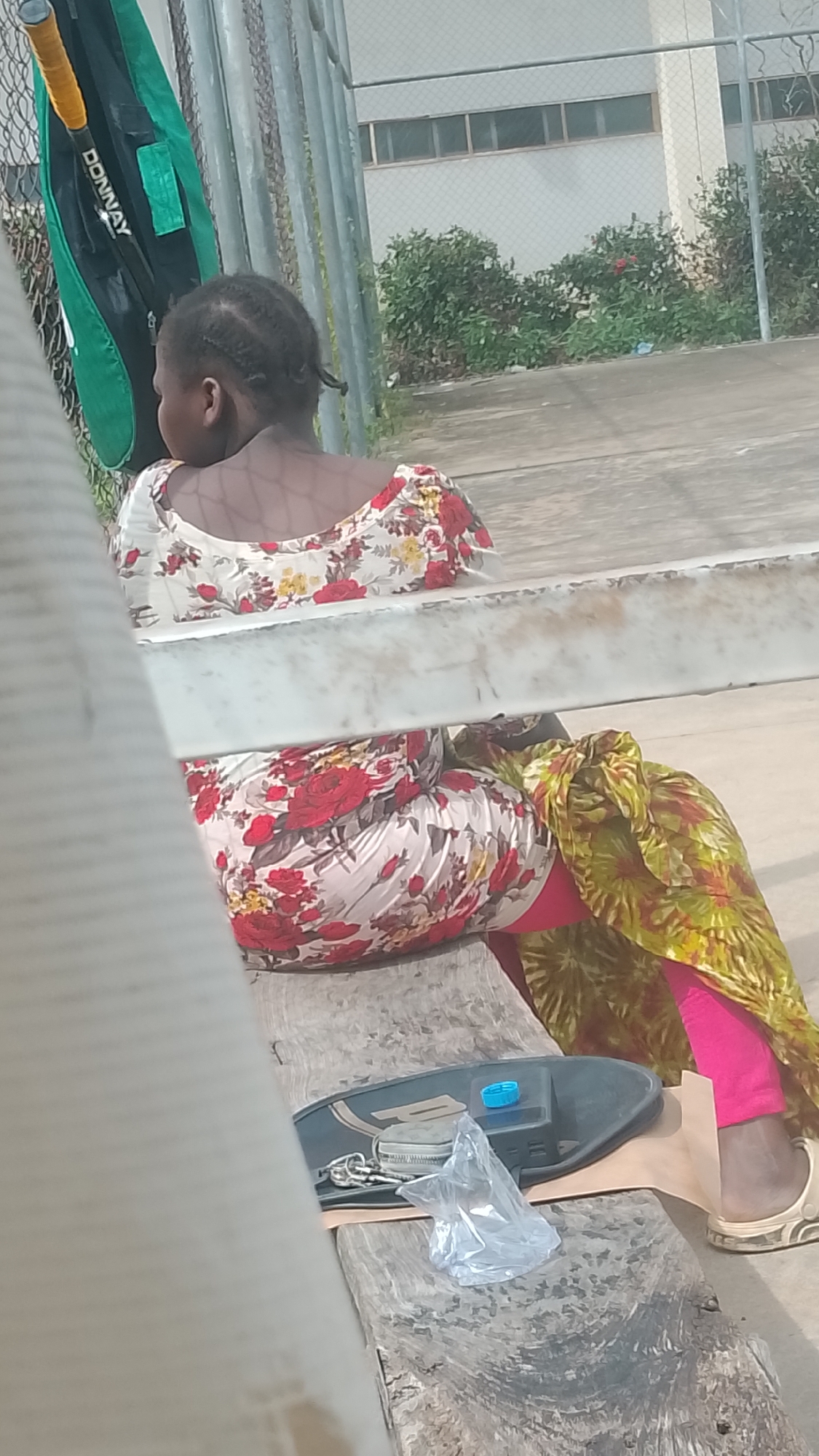 The image displays an outdoor scene located in Lokoja, Nigeria, featuring a seated individual and surrounding environmental elements. In the foreground, a person, appearing to be a woman with dark skin and dark hair styled in cornrows, is seated on a weathered wooden bench, viewed from behind. She wears a white top and a skirt, both adorned with a red and yellow floral pattern. Bright pink leggings are visible beneath a green and yellow patterned fabric wrap or scarf draped over her legs. Her right foot, clad in a light-colored open-toed sandal, rests on the ground.
On the wooden bench to her right, a dark-colored, possibly plastic, tray or container holds a blue plastic bottle cap, a set of keys on a ring, and a crumpled clear plastic bag. A piece of brown paper or cardboard is partially visible beneath these items.
To the left of the seated individual, a green tennis racket bag with "DONNAY" printed in white letters is visible, likely hanging on a metal fence. A yellow racket grip protrudes from the bag. The background features a chain-link fence with vertical metal posts. Beyond the fence, a light-colored building with multiple horizontal rectangular windows is visible. Green bushes and some red flowering plants grow in front of the building. The ground is a concrete or paved surface.