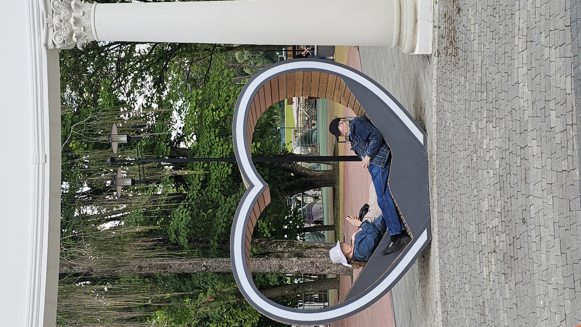 An elderly couple is relaxing on a heart-shaped bench in a park, possibly in the city of Alikonovka, Russia. The man, wearing a black baseball cap and a denim jacket, is lying down with his eyes closed. The woman, wearing a white sun hat and a denim jacket, is sitting up and looking at her phone. The bench is made of wood and metal and is situated on a paved area with trees and streetlights in the background. A large white column is visible on the right side of the image.
